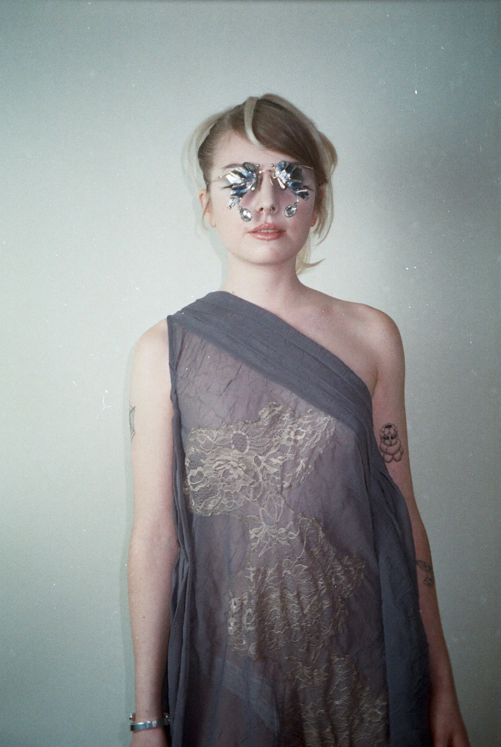 Joven con gafas decorativas de espejo, vestido gris con encaje, y tatuajes en brazos, posa en un fondo simple.