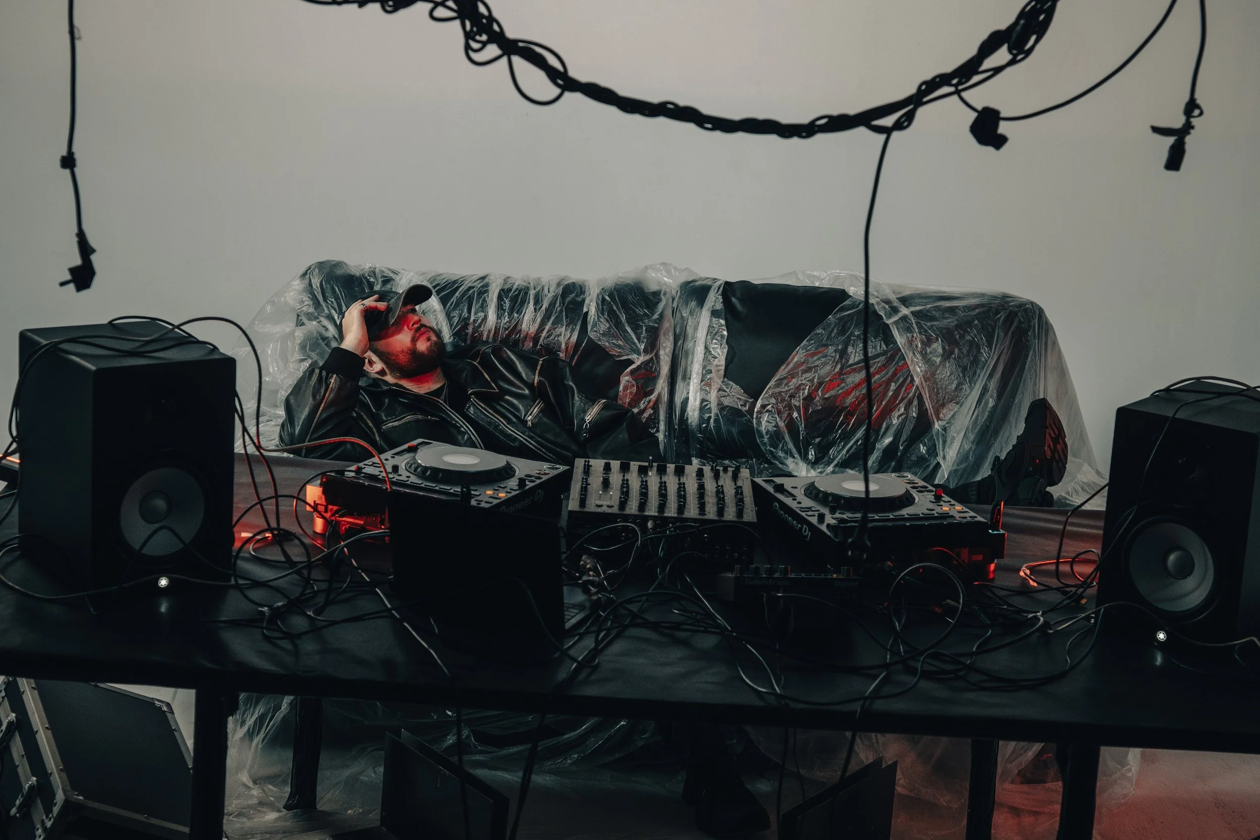 Un hombre con camiseta negra y gorra descansando en un sofá cubierto con plástico, rodeado de equipo de DJ y altavoces en un ambiente oscuro con cables colgando del techo.