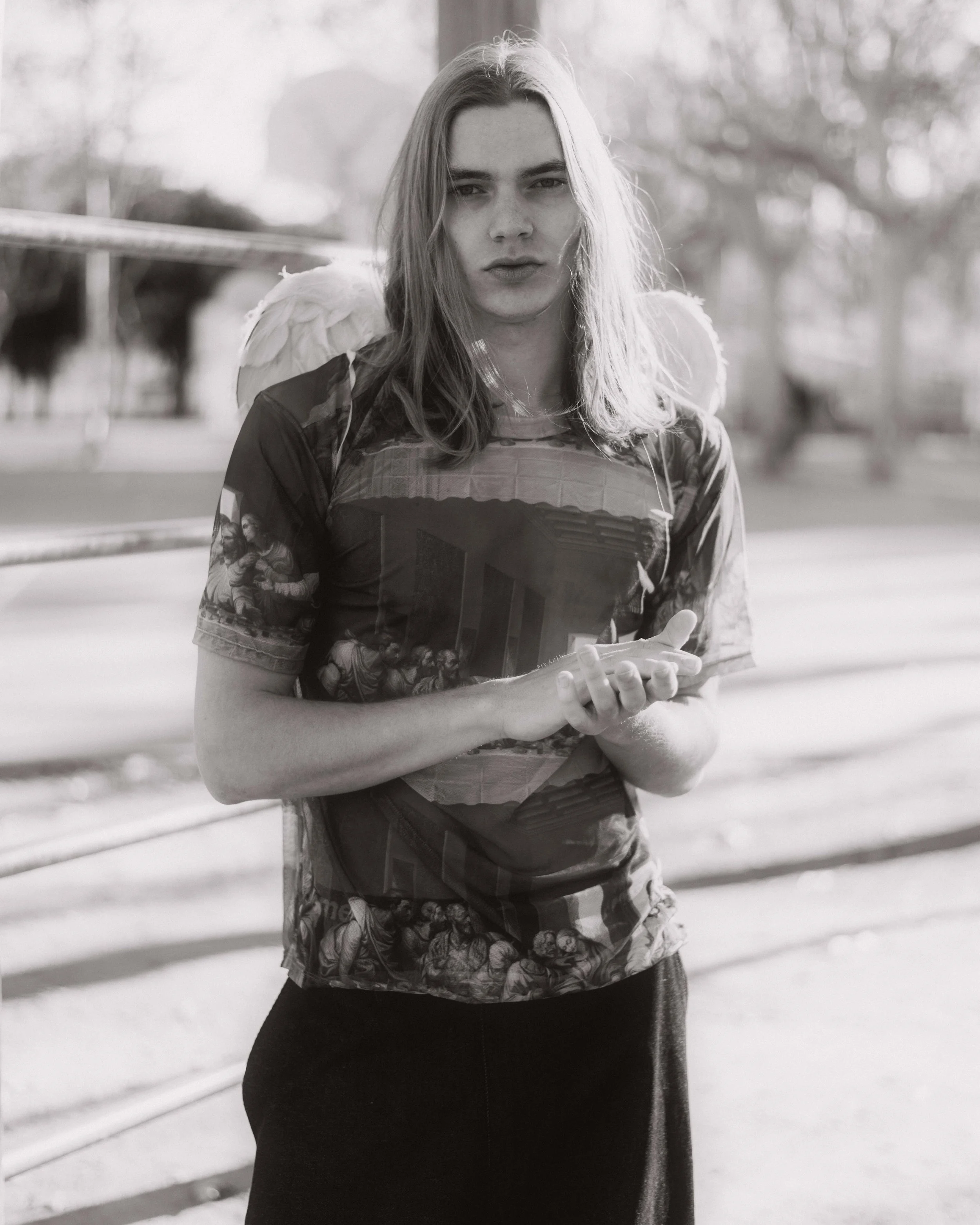 Joven mujer con cabello largo y rubio, con alas de ángel, de pie al aire libre, usando una camiseta con impresión y sosteniendo un teléfono móvil, en un entorno natural con árboles y una estructura metálica en el fondo.