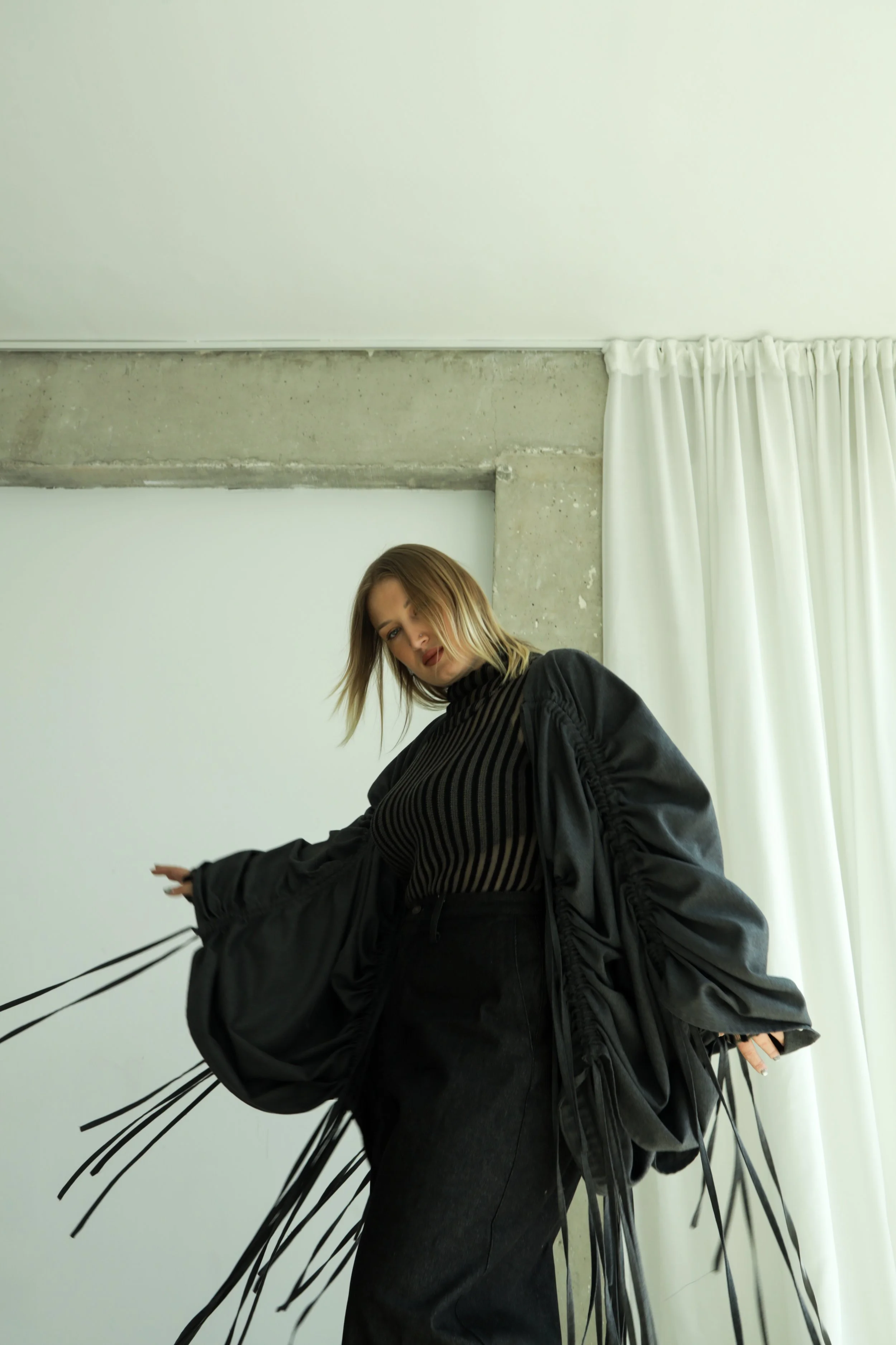 Mujer con cabello rubio, vestido negro y abrigo negro con cordones largos, posando frente a la pared con cortinas blancas.