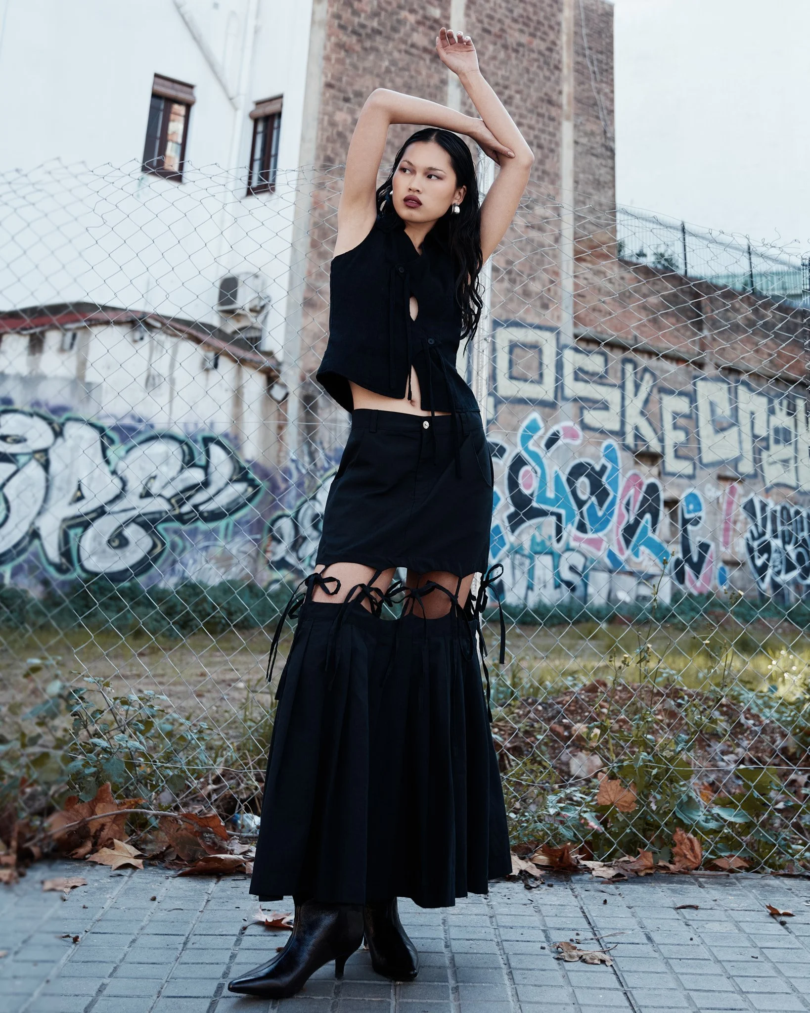 Mujer con vestido y botas negras posando frente a una cerca de alambre y graffiti en la pared, en una escena urbana