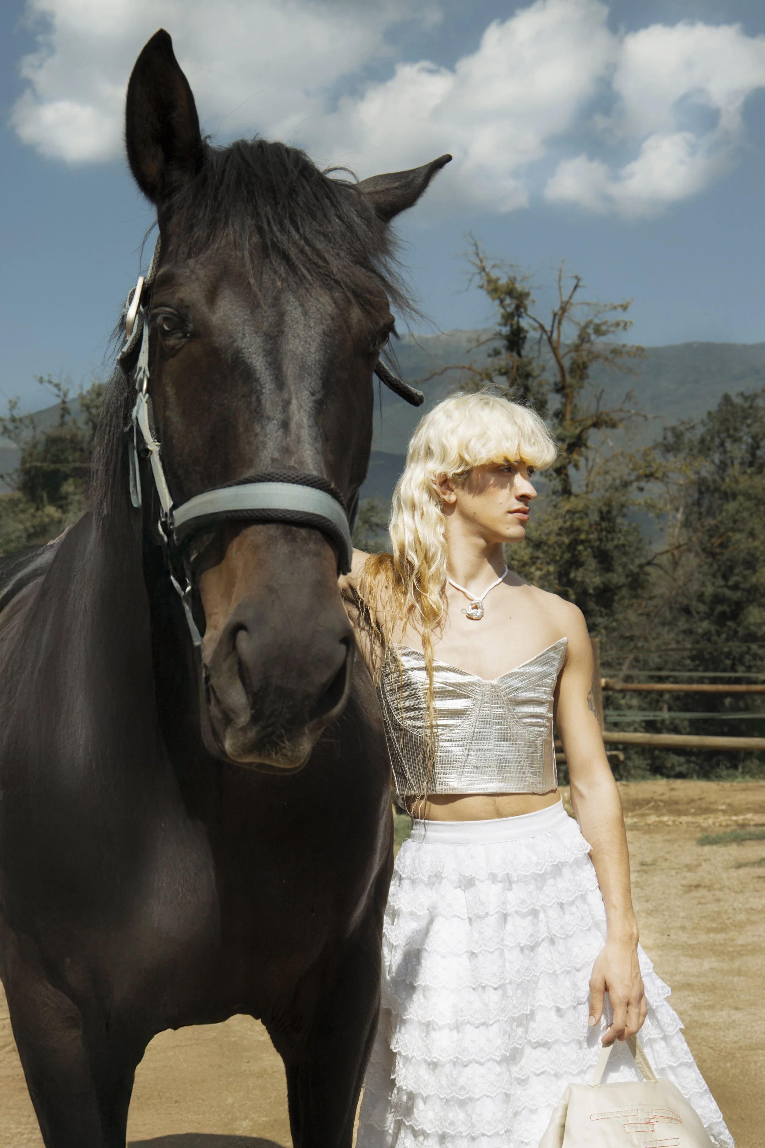 Una persona con cabello rubio y largo, vestida con un top plateado y una falda blanca, junto a un caballo negro en un campo al aire libre con árboles y montañas de fondo.