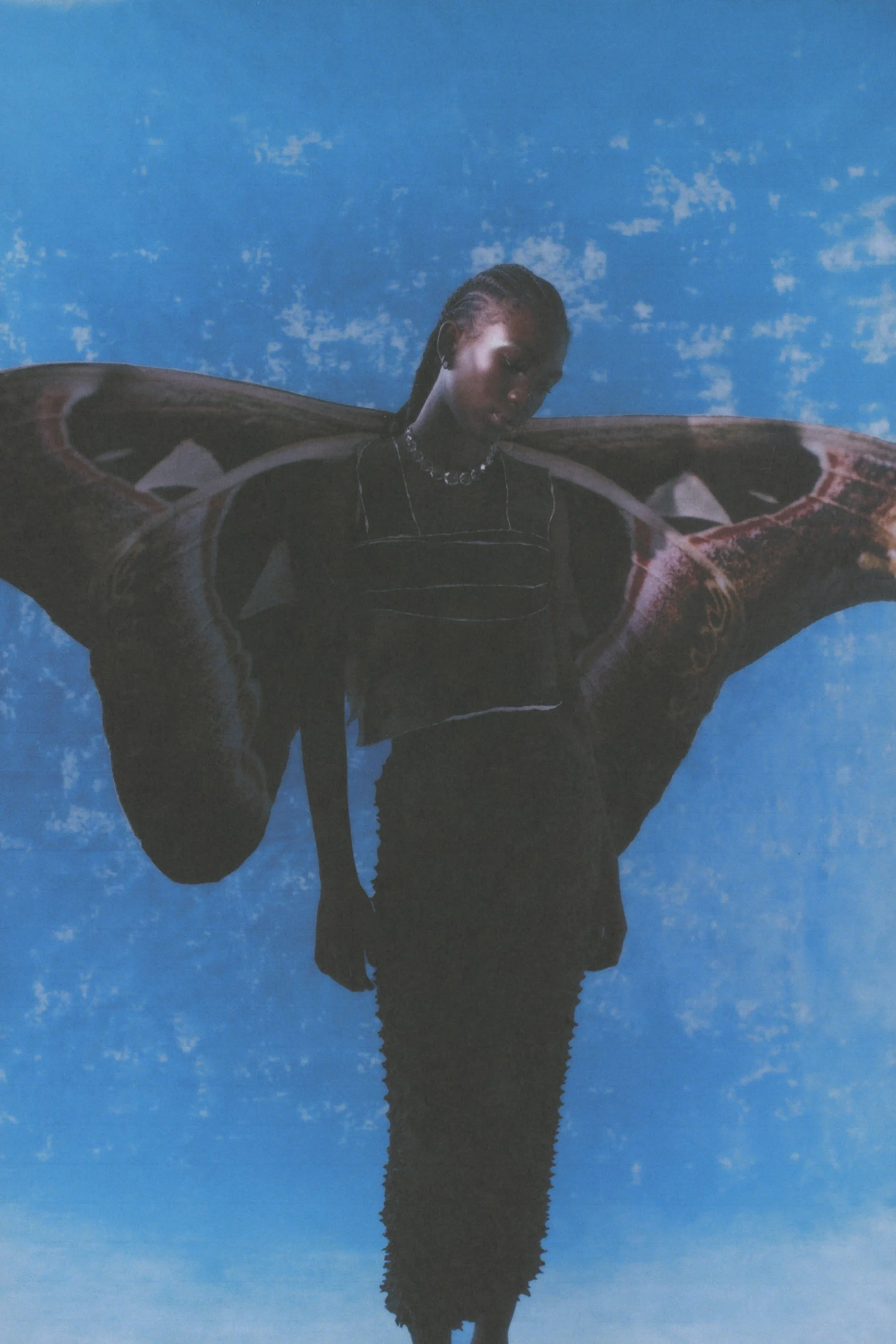 Una persona de cabello trenzado, vestida con ropa negra, sostiene una gran figura de mariposa en el aire con fondo azul con nubes.