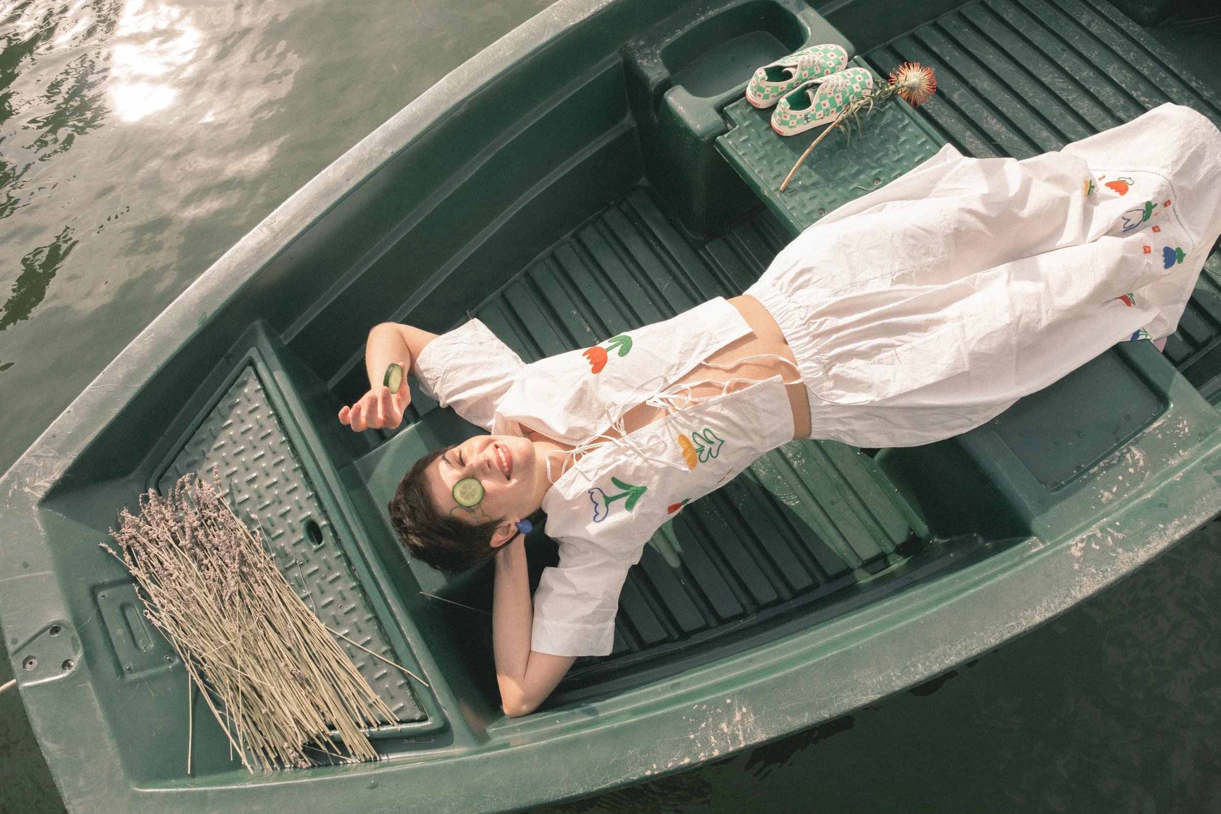 Mujer descansando en una embarcación con rodajas de pepino en los ojos, rodeada de objetos como sandalias con estampado floral, cabello suelto, y con un fondo de agua