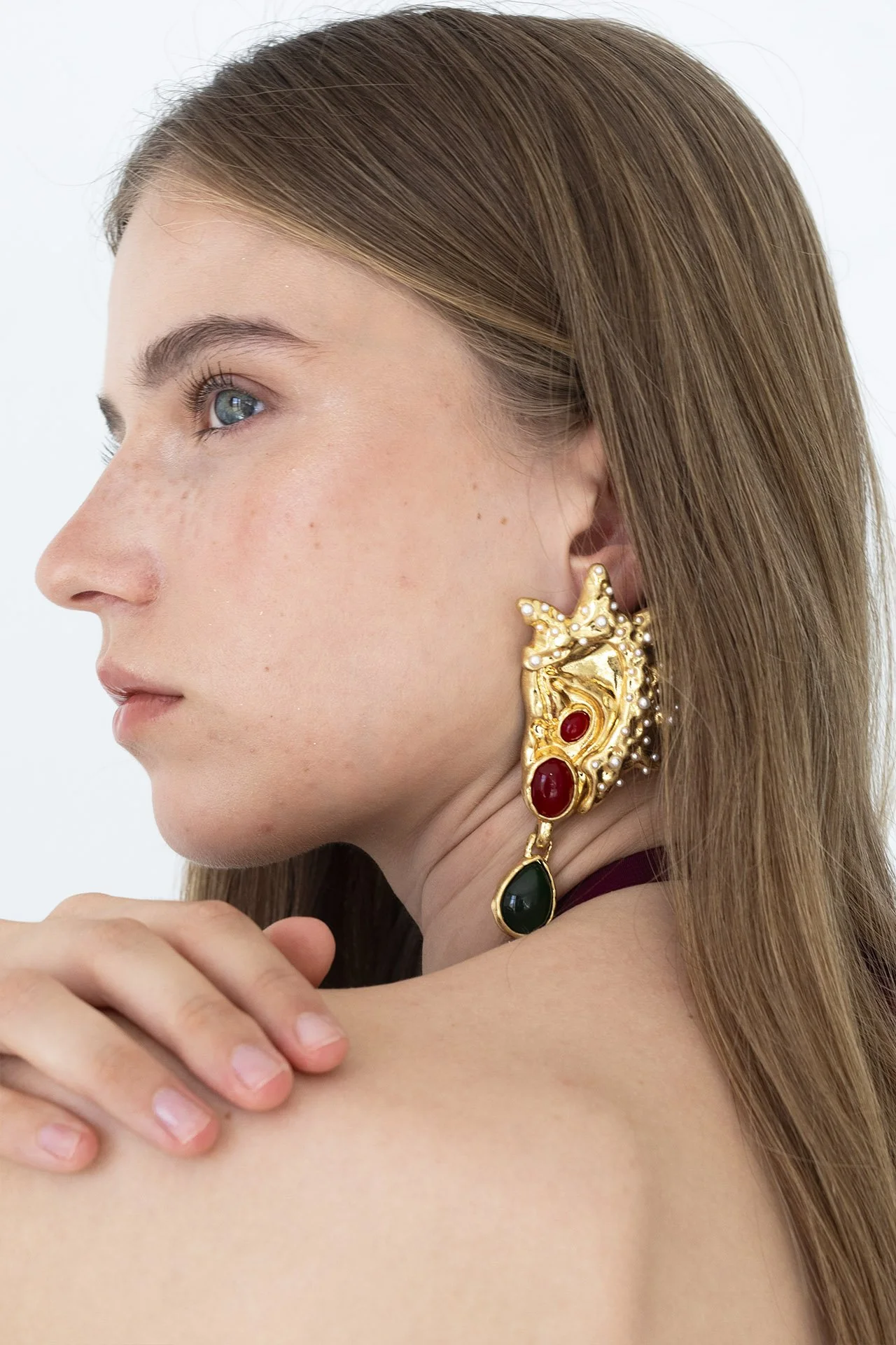 Mujer con cabello castaño, piel clara, usando un pendiente dorado con perlas, rubíes y un dije de piedra verde, tocándose el hombro izquierdo.