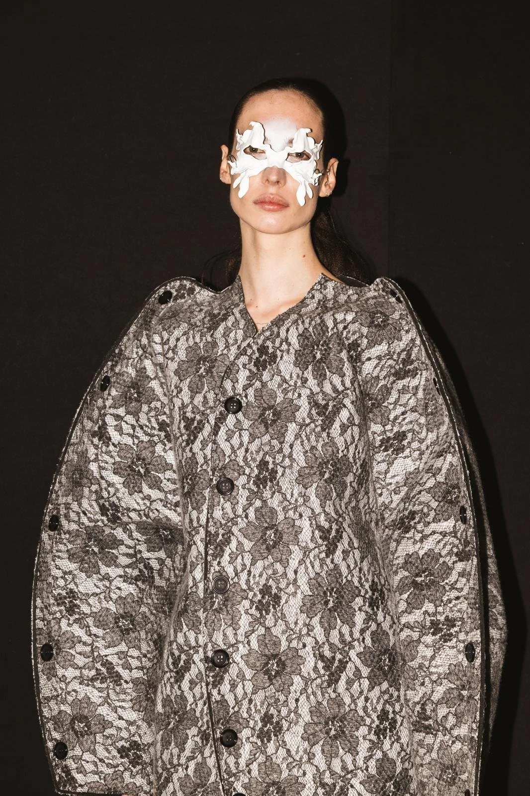 Una mujer con maquillaje de papel en el rostro, con cabello oscuro, lleva puesto un abrigo con estampado de encaje en tonos grises y marrones, contra un fondo negro.