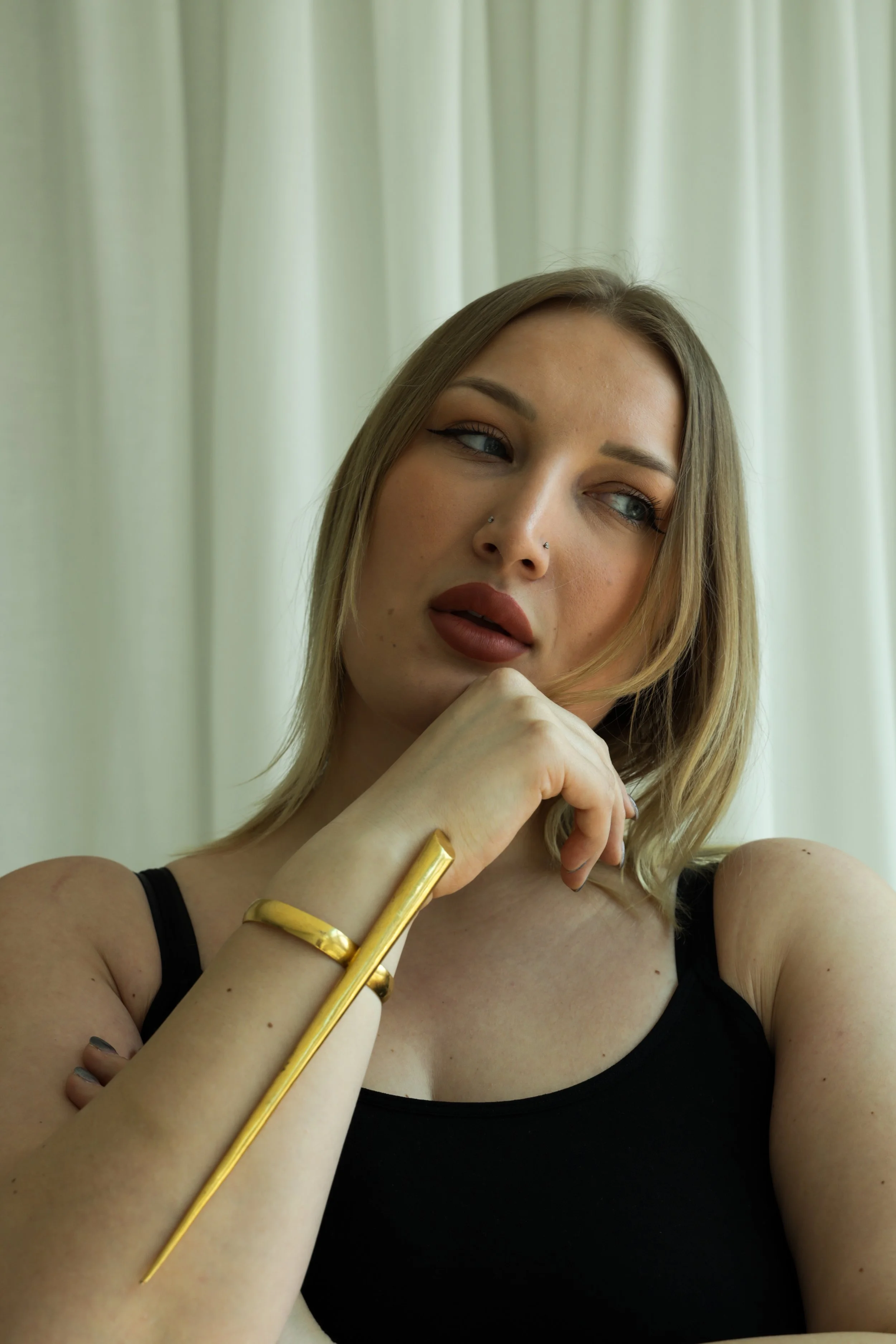 Joven mujer con maquillaje y labial marrón, con pendientes de argolla pequeños, usando una blusa negra, posando con su mano en el rostro, usando un brazalete y un pincho dorado en la muñeca, contra cortinas blancas.