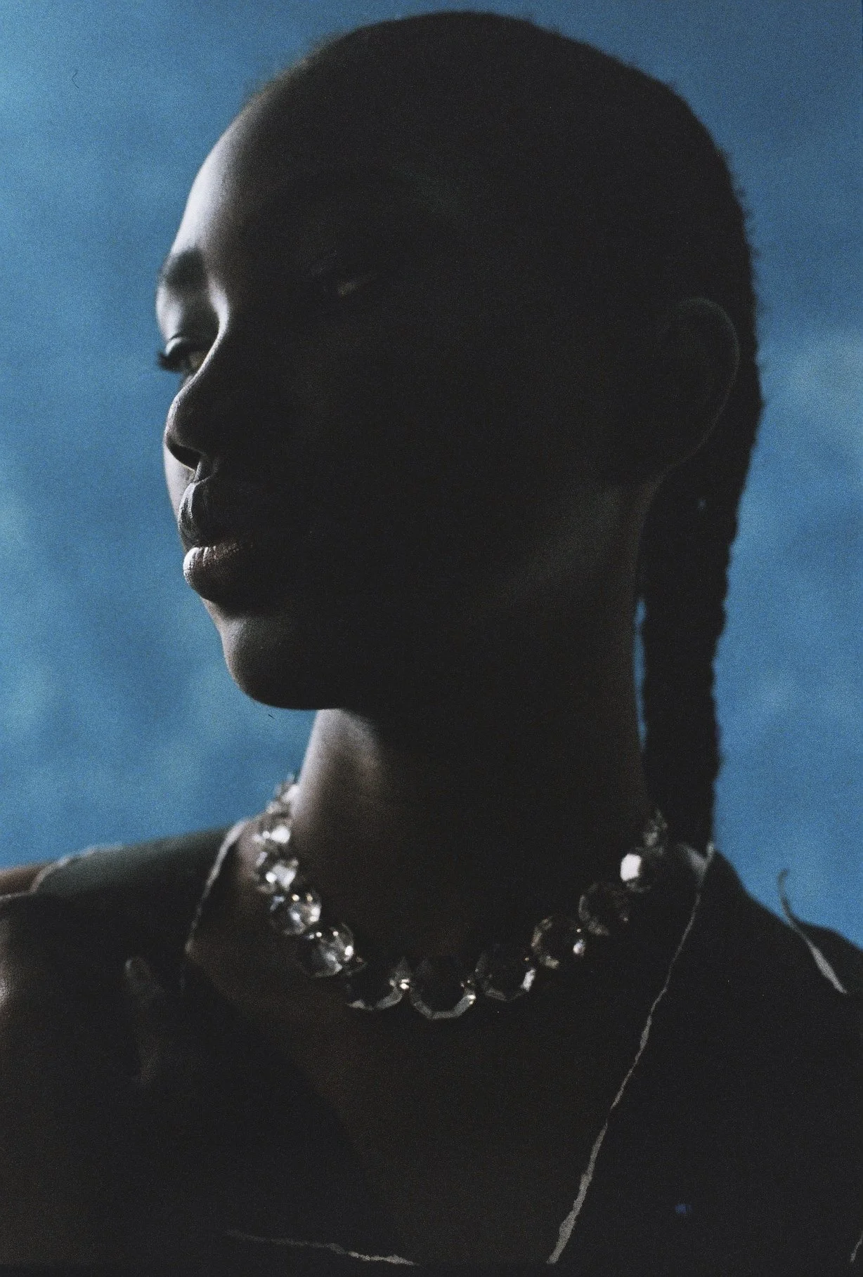 Retrato de una mujer con cabello trenzado, usando un collar de piedras grandes y un arete, con una expresión pensativa, en un fondo azul.