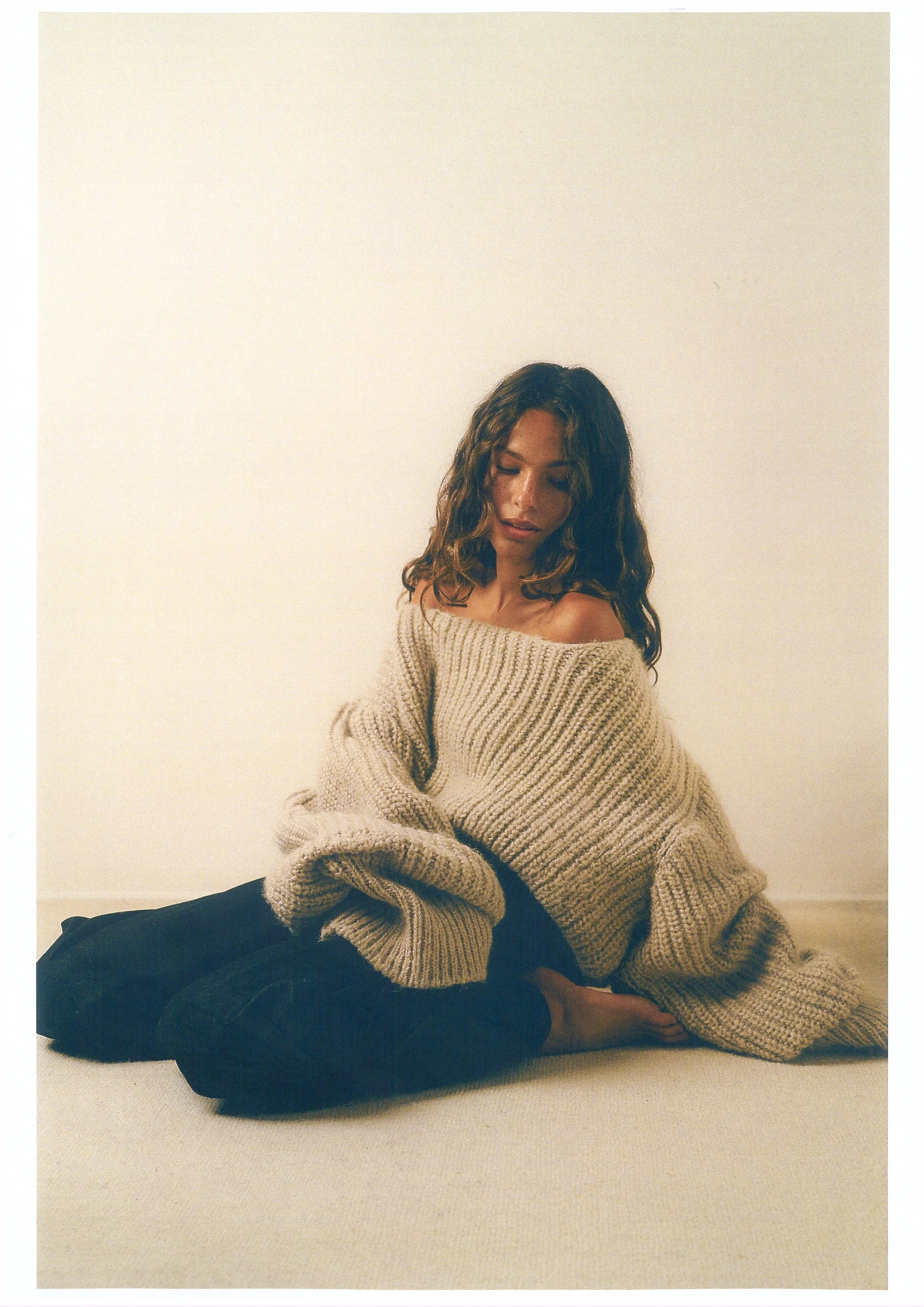 Mujer con cabello rizado, usando un suéter gris y pantalones negros, sentada en el piso junto a una pared blanca, con expresión pensativa.