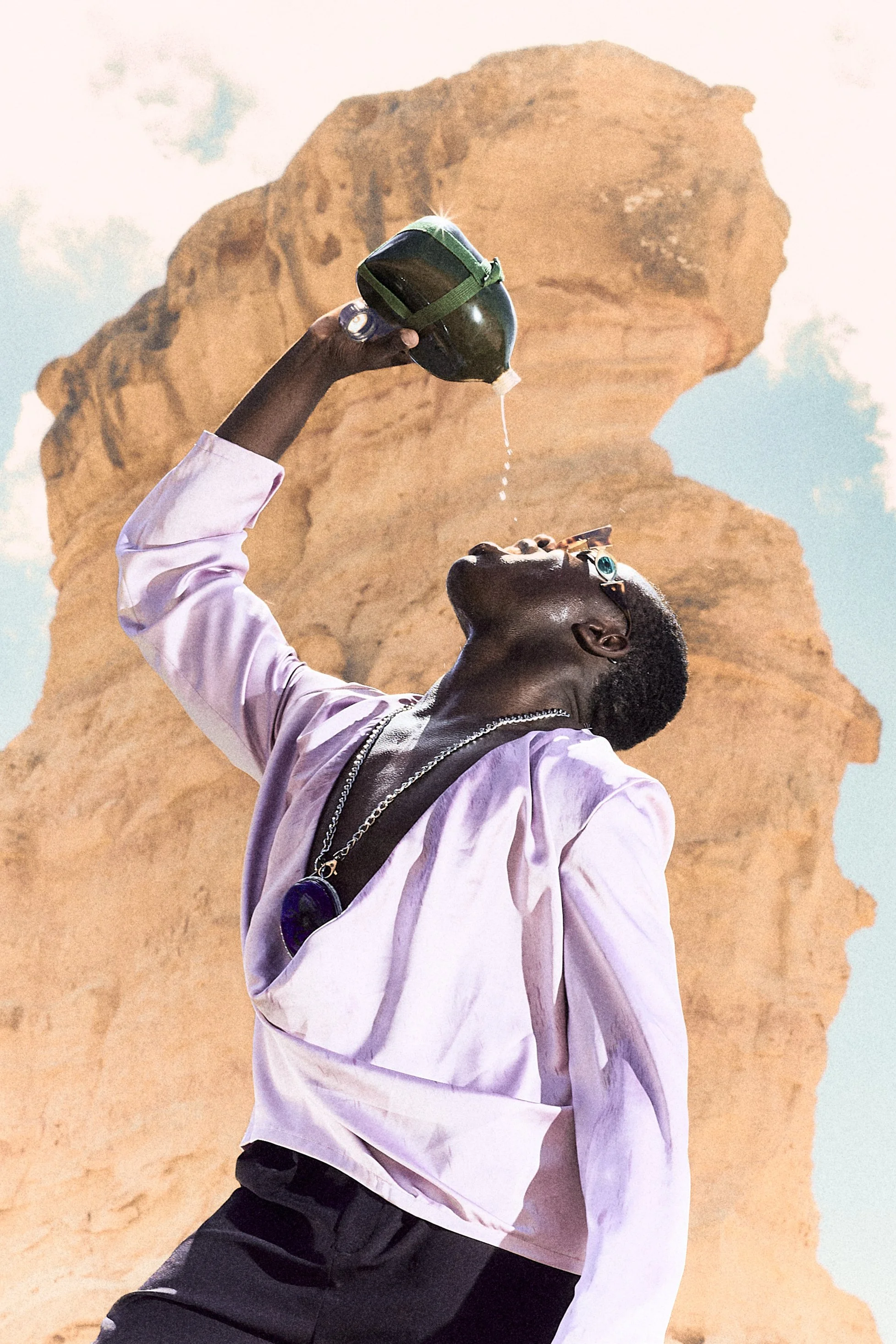 Una persona con gafas, camisa rosa y collares, inclinada hacia atrás, vertiendo agua de una cantimplora en su boca, con una formación rocosa en el fondo y cielo con nubes.