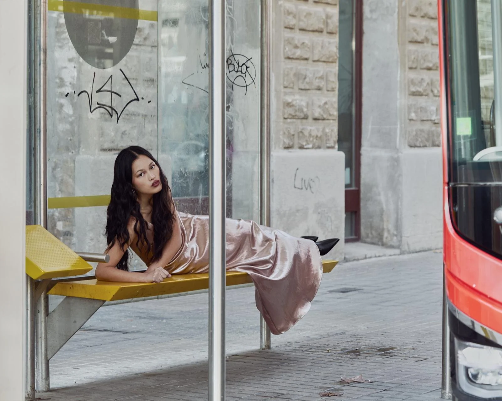 Mujer con vestido de seda acostada en una banca de autobús, en un paradero urbano, con paredes de ladrillo y graffiti en el fondo.