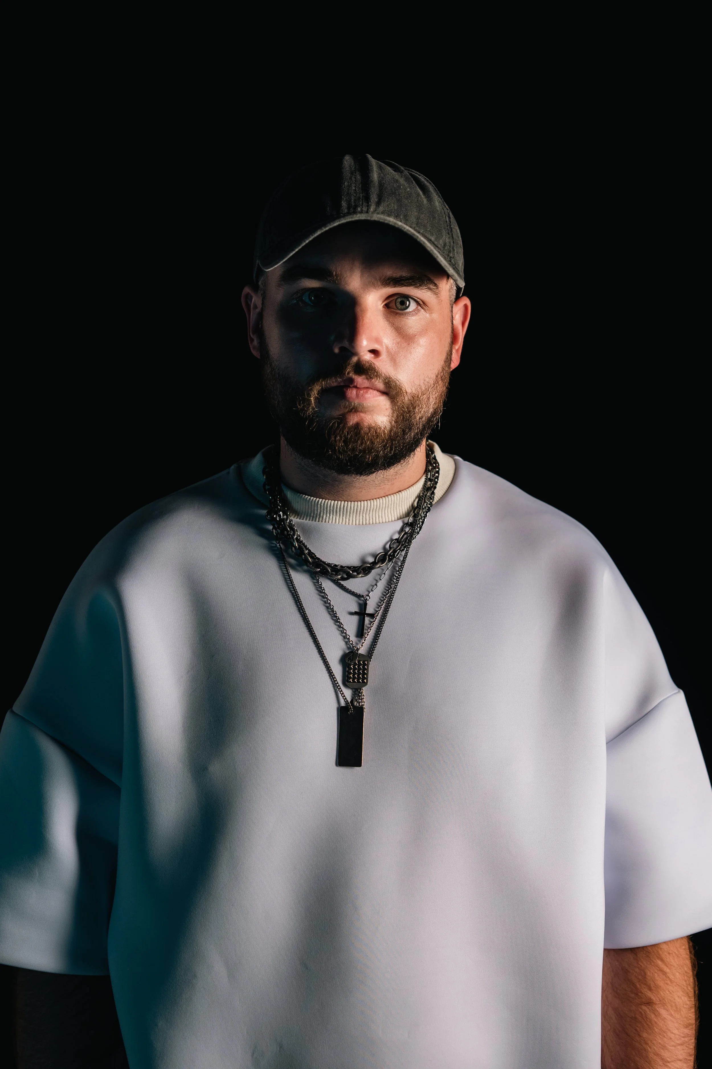 Retrato de un hombre con barba, usando gorra, camisa blanca y varias cadenas, frente a fondo negro con iluminación lateral.