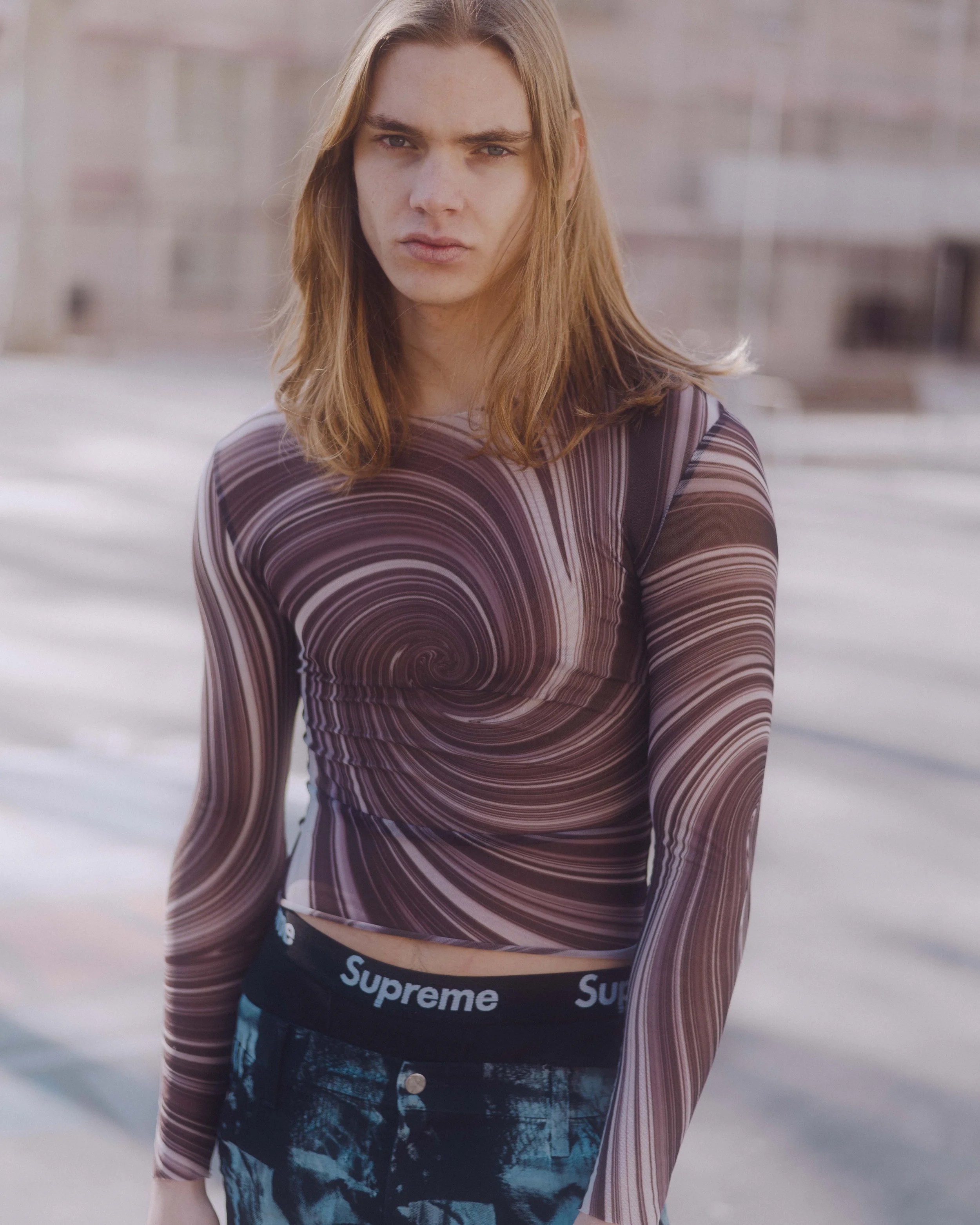 Joven con cabello largo y rubio, viste una camiseta de manga larga con patrón en espiral y pantalones con banda de marca 'Supreme', en un entorno urbano.