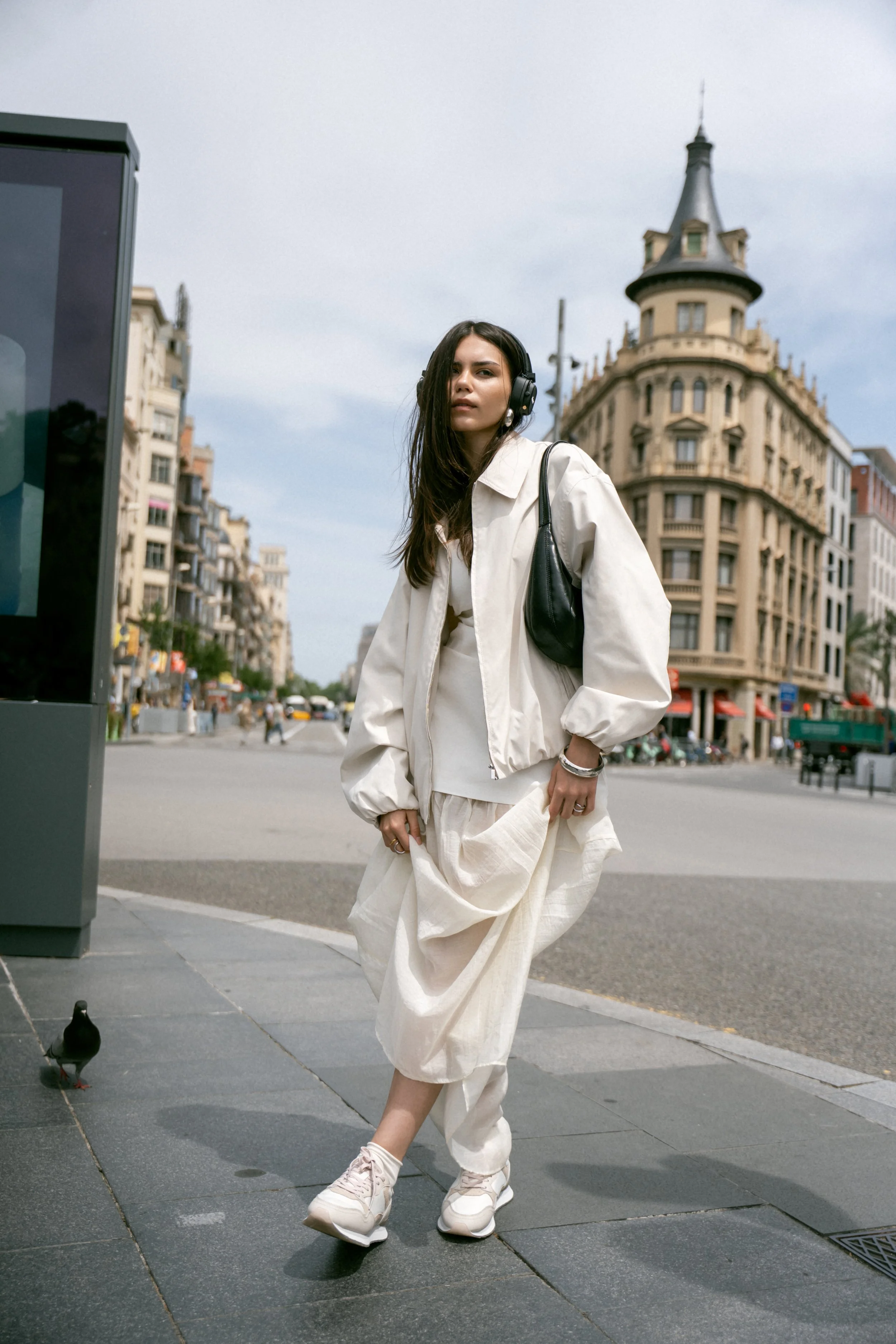Mujer caminando en la ciudad con auriculares, vestida con ropa blanca y bolso negro, en una acera urbana junto a una Pidgeon en la calle.