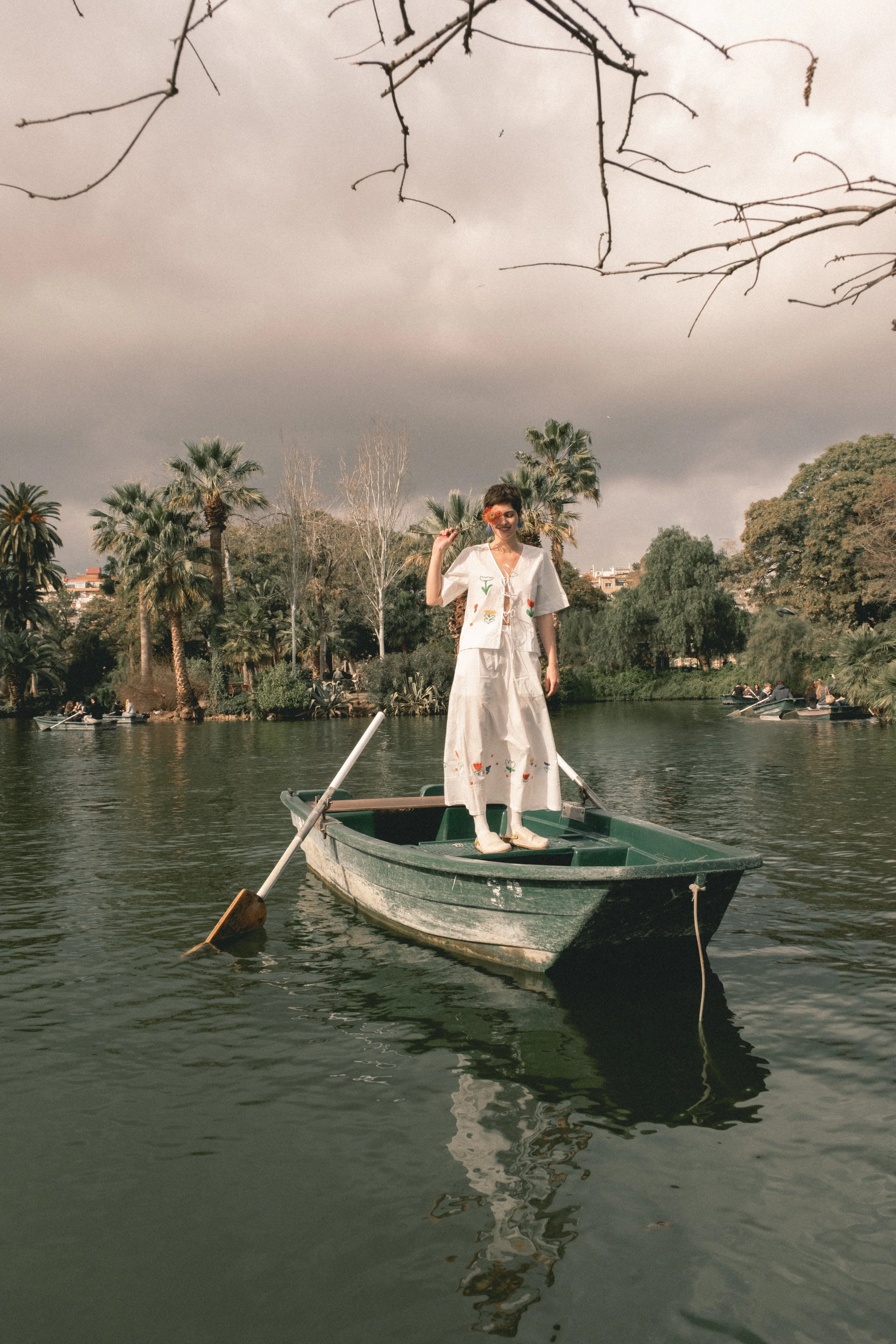 Persona vestido con ropa blanca decorada con dibujos de flores y frutas, de pie en un bote en medio de un lago en un parque con palmeras, con un cielo nublado.