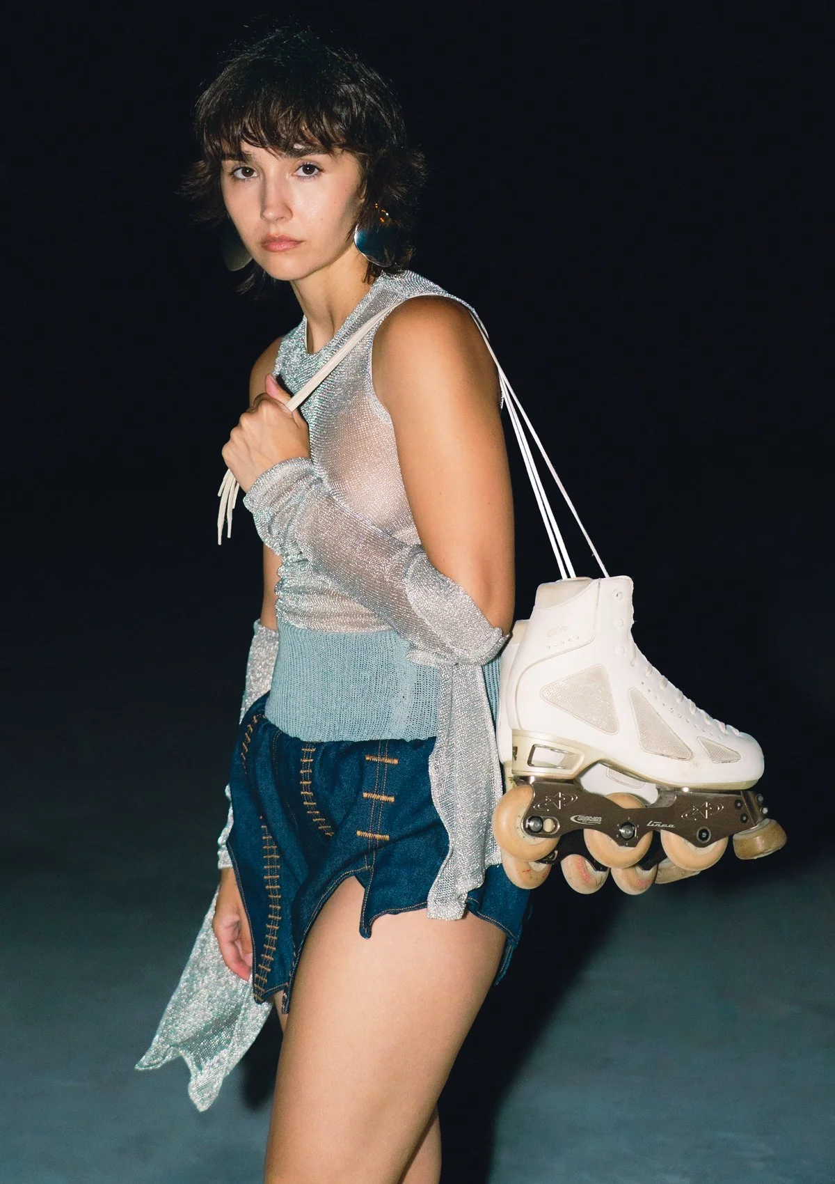 Mujer con ropas modernas y cortas, sujetando patines en línea blancos con una mano, en un fondo oscuro.