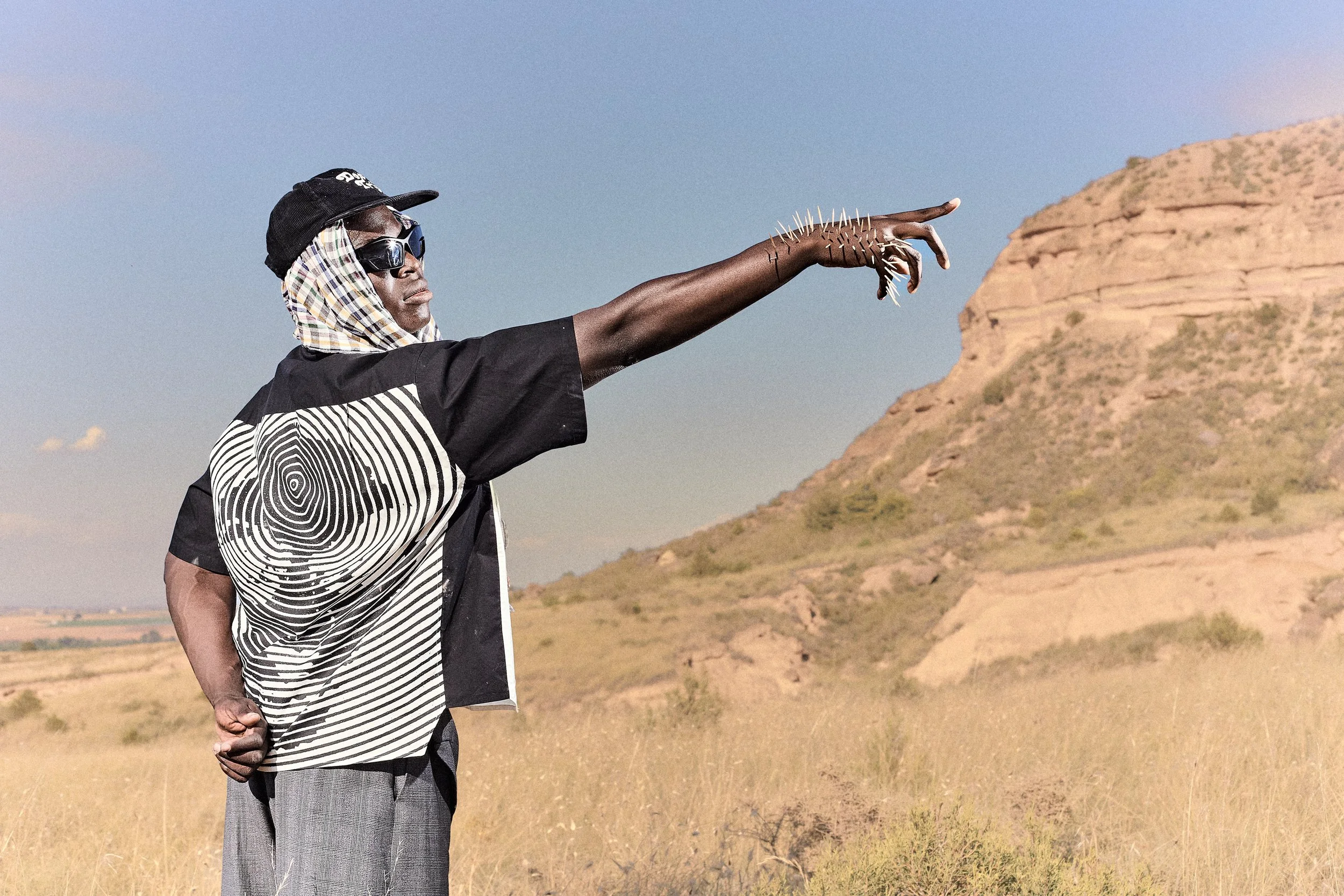 Un hombre de piel oscura vestido con ropa de estilo artístico y accesorios en un paisaje desértico, señalando hacia el horizonte con la mano extendida.