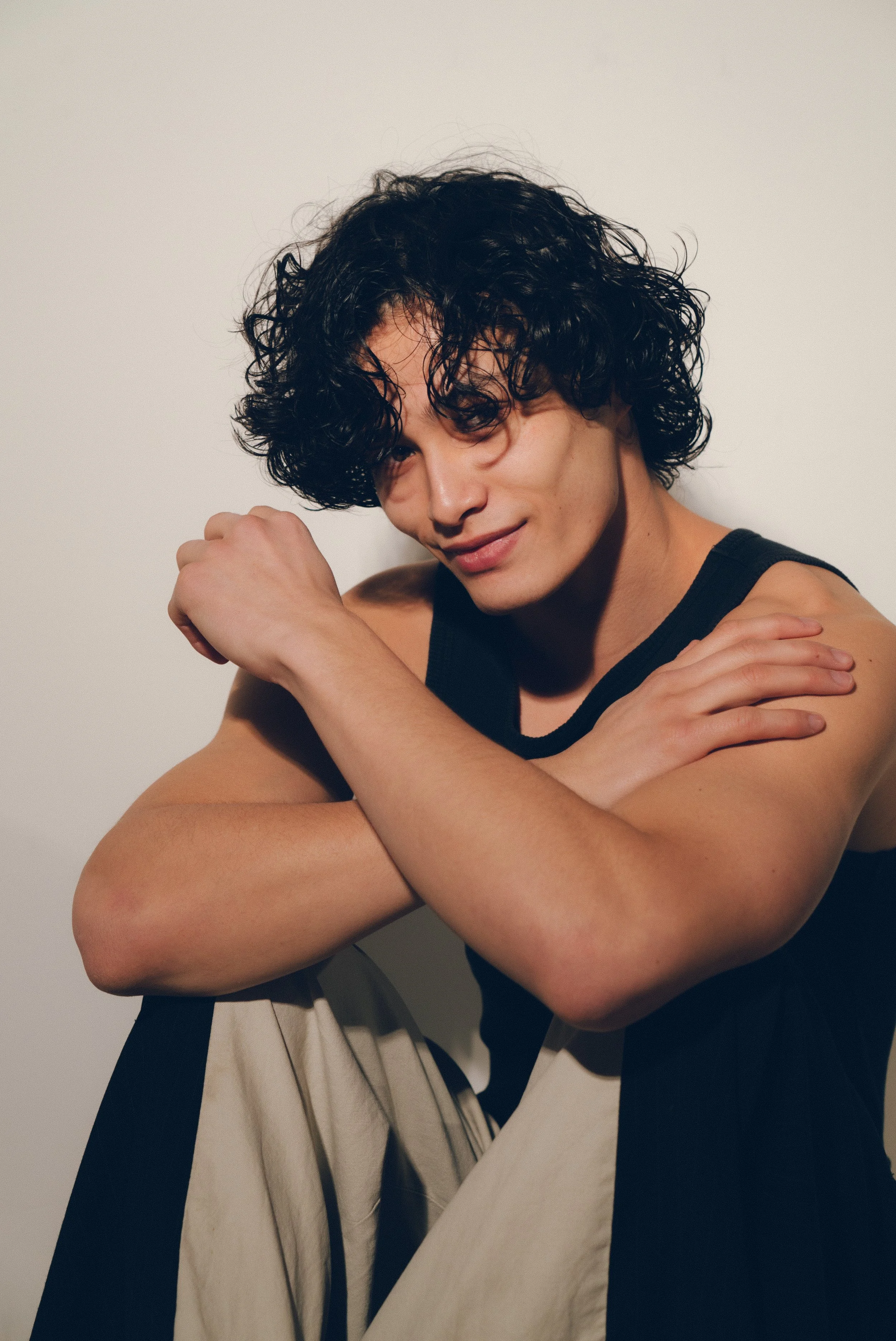 Joven con cabello rizado, piel clara, vestido sin mangas negro y pantalones claras, sentado en el suelo frente a una pared blanca, mirando con una expresión relajada y sonriendo levemente.