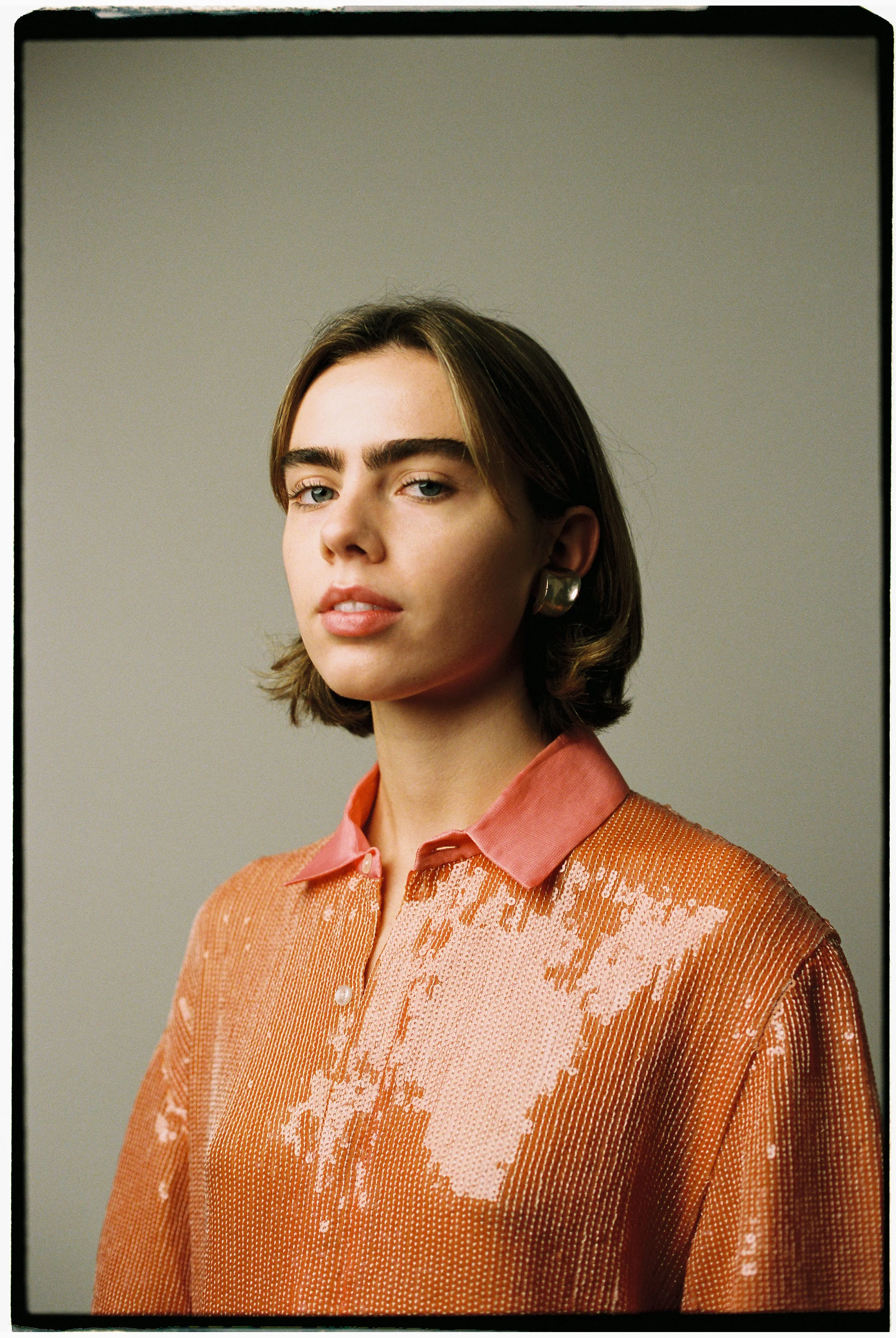 Retrato de una joven con cabello castaño, cejas espesas y expresión segura, usando blusa de color coral con detalles brillantes y un par de aretes grandes, contra un fondo gris claro.
