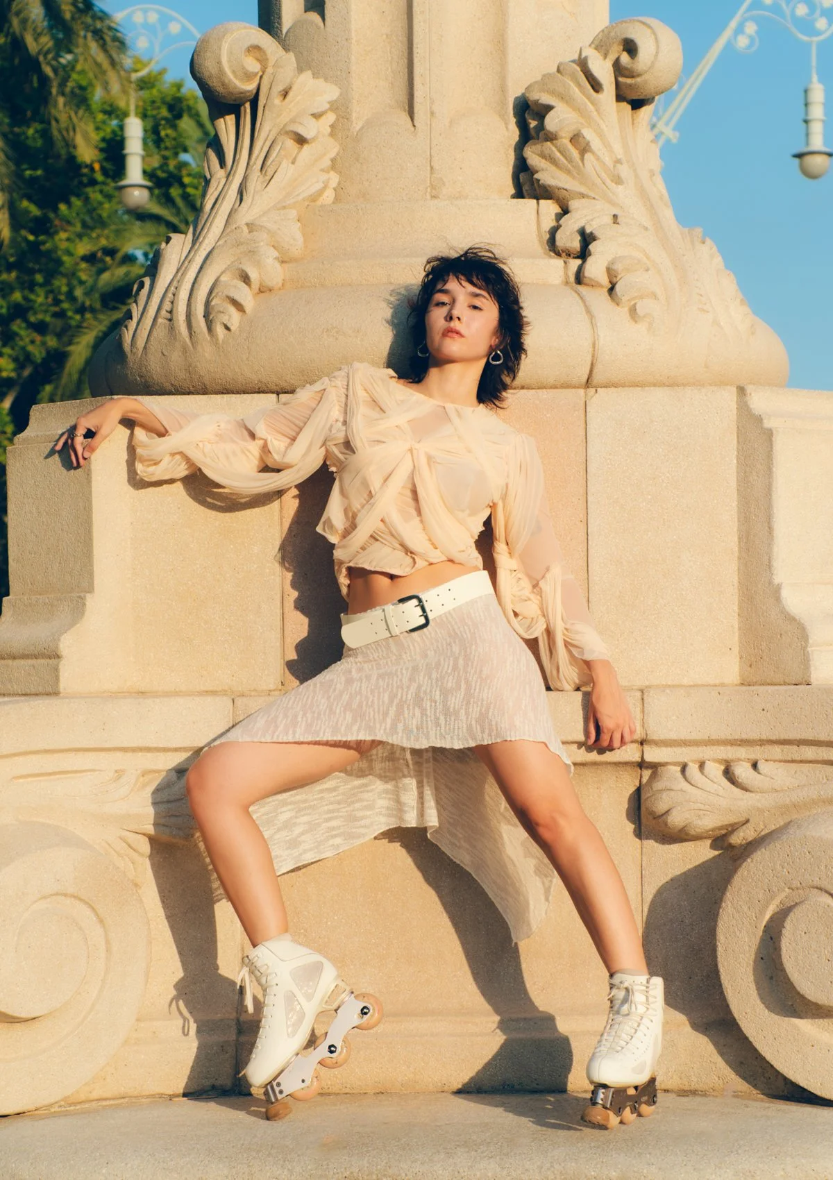Joven mujer posando con patines sobre ruedas frente a una fuente de piedra en un parque, vestida con ropa beige y estilos modernos.