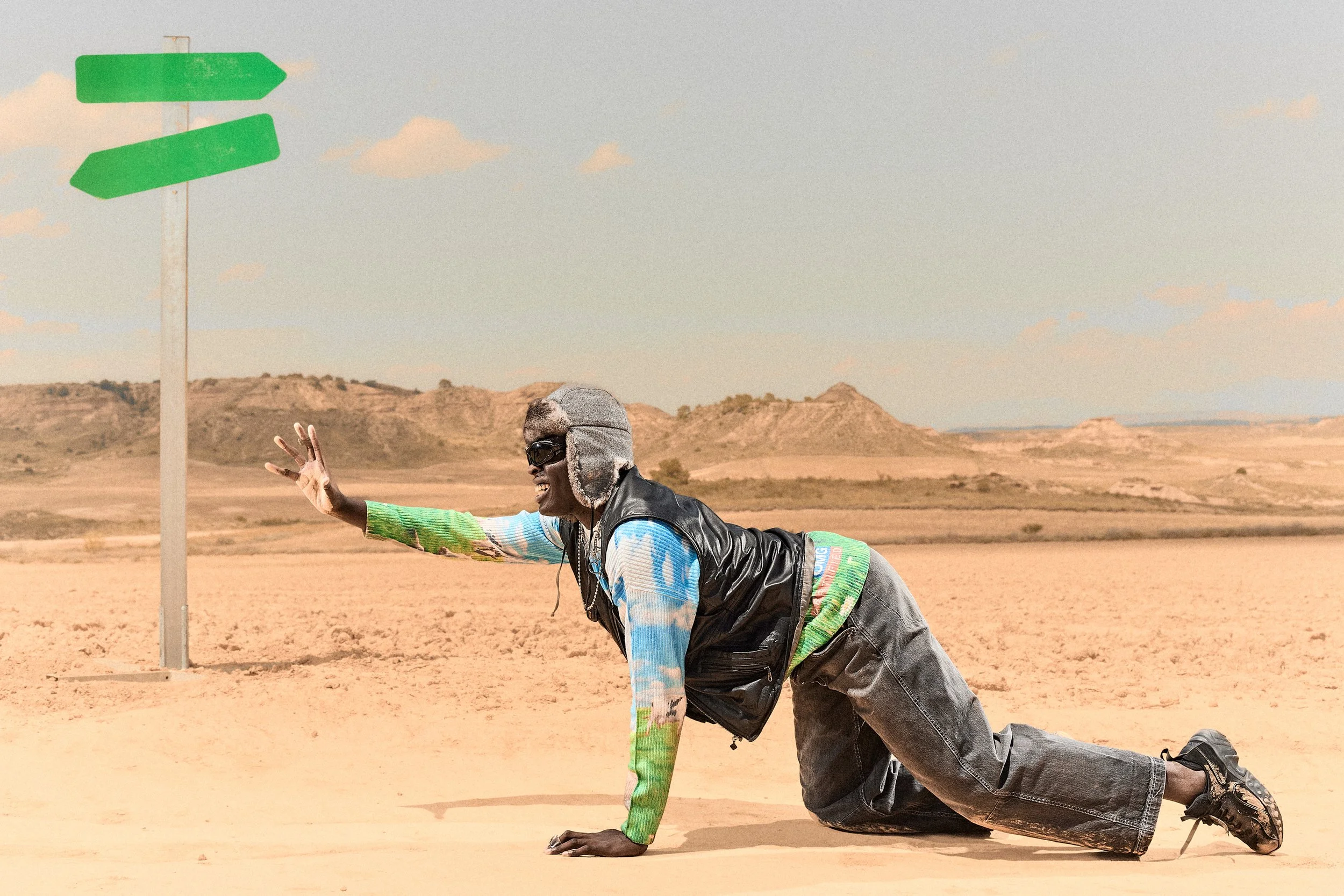 Hombre en estado de agonía en medio del desierto, usando ropa colorida, gorro con orejeras y gafas de sol, con un señalamiento verde en el fondo.