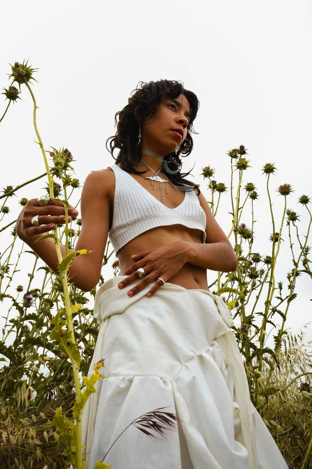 Mujer joven con ropa blanca en un campo de plantas altas, con un cielo nublado de fondo.