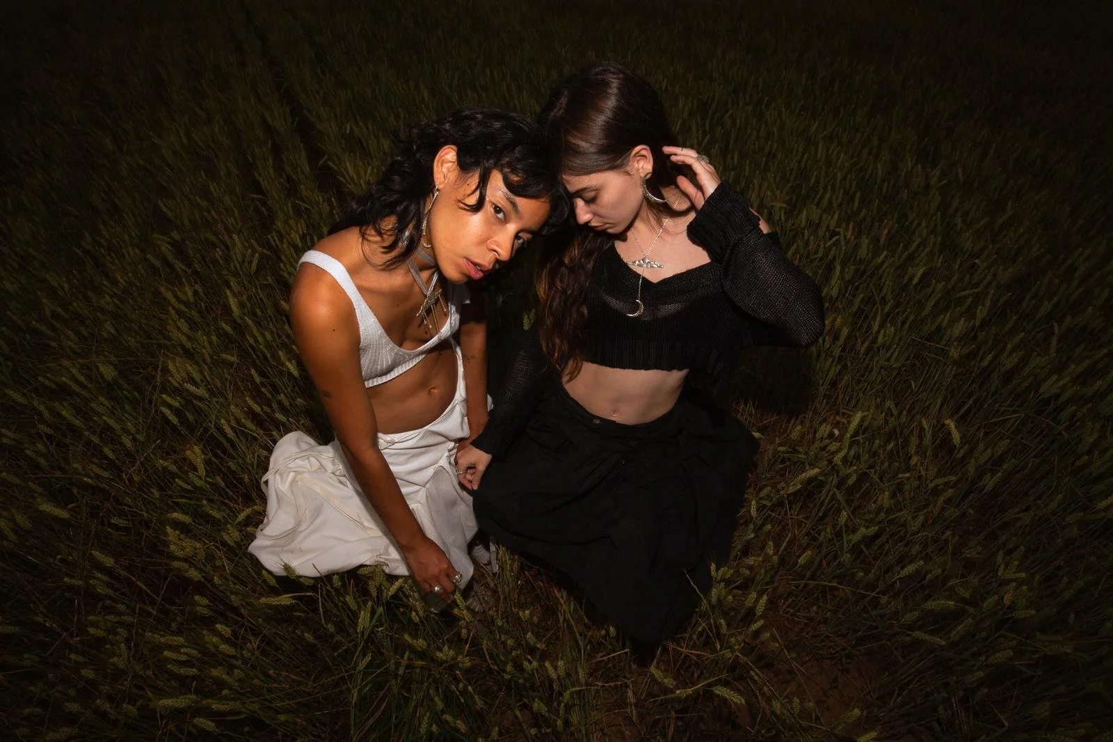 Dos mujeres jóvenes en un campo de hierba alta, una vestida con ropa blanca y la otra con ropa negra, en una pose artística y expresiva.