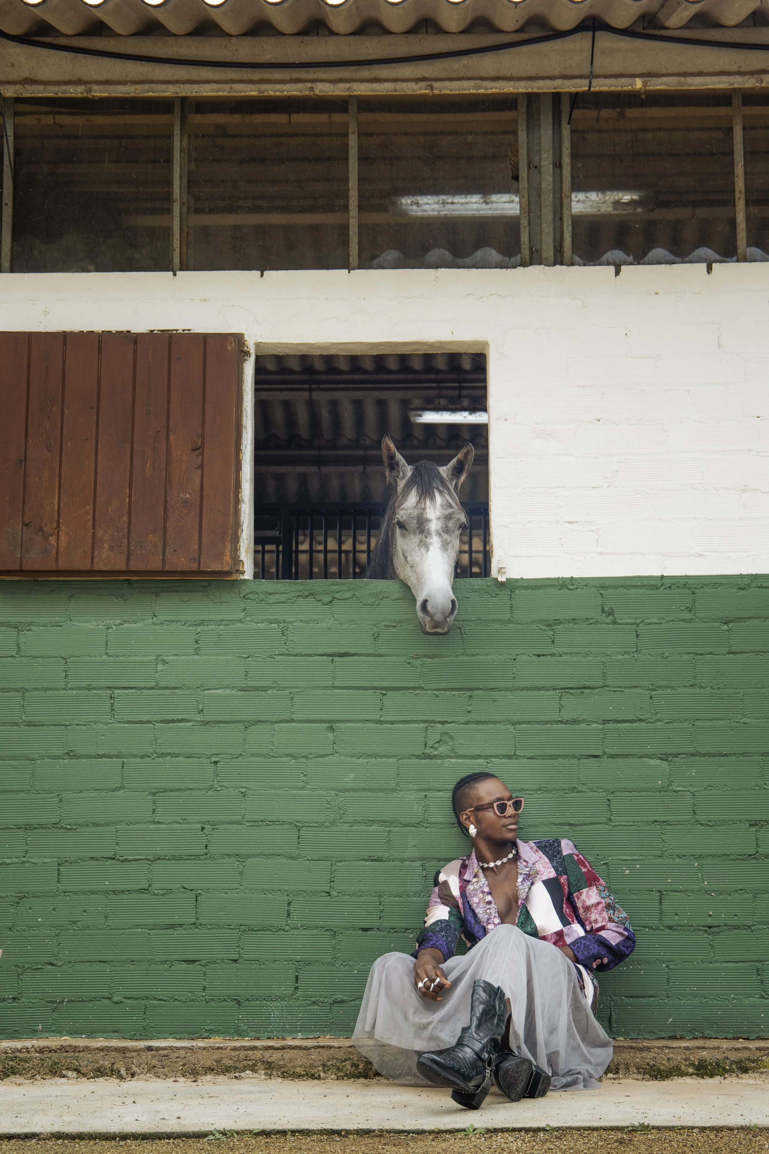 Una mujer sentada con estilo frente a un muro verde, usando gafas y joyas, y un caballo asomándose desde una ventana superior.