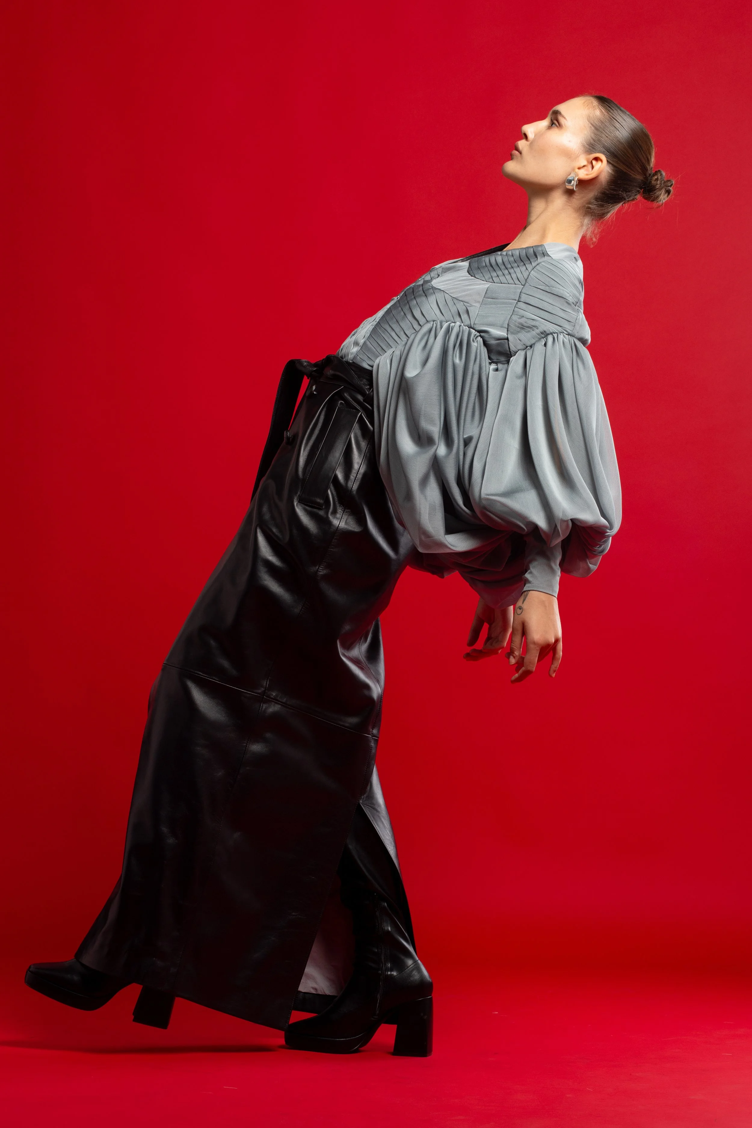 Mujer de pie con vestido gris con mangas abullonadas y falda negra de cuero, de perfil contra fondo rojo.