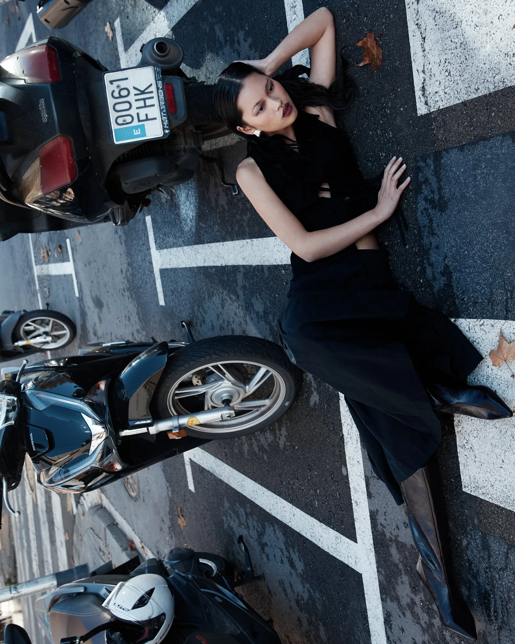 Mujer con cabello negro y vestido negro acostada en un estacionamiento, rodeada de motocicletas, con una hoja caída a su lado.