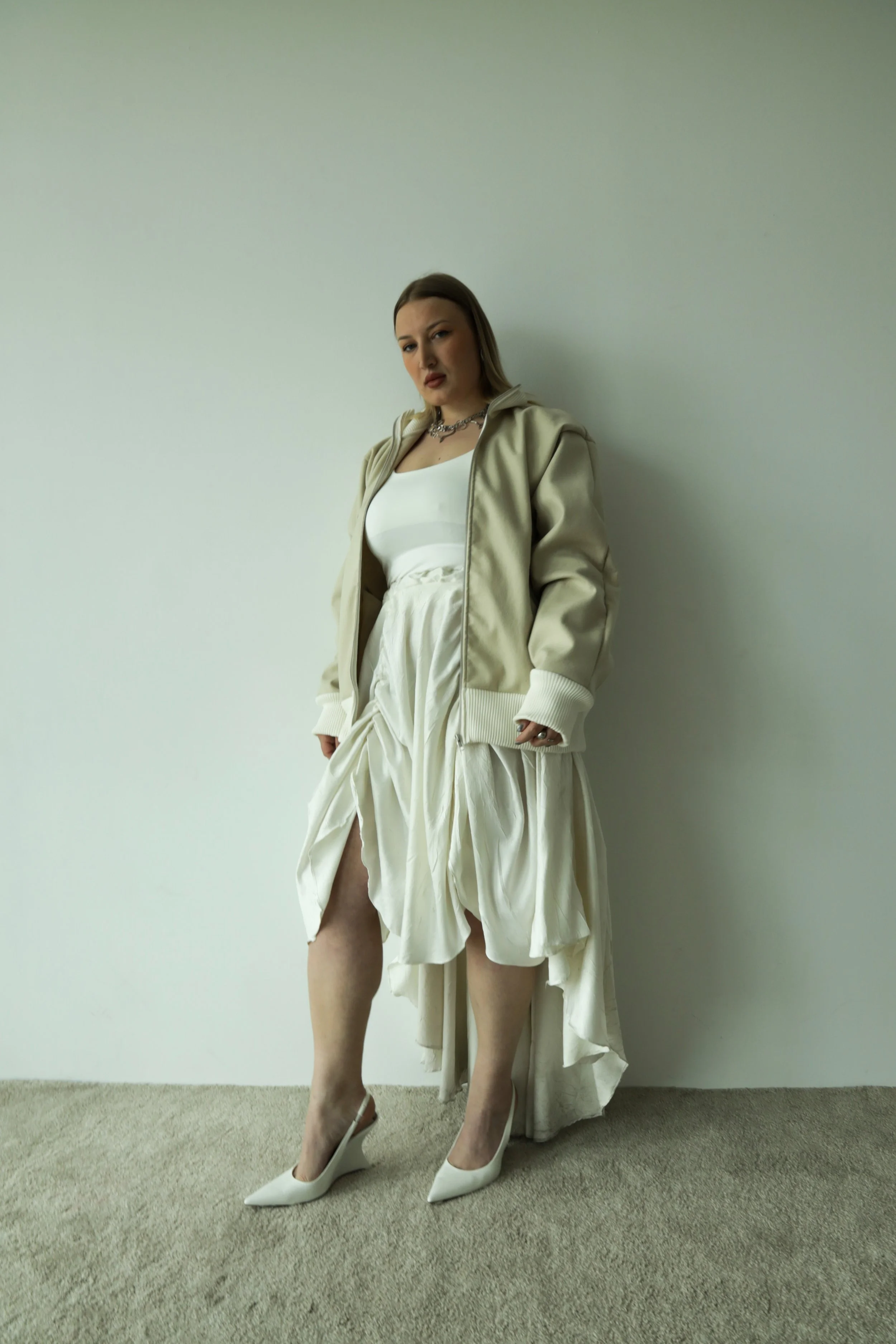 Mujer con ropa blanca y una chaqueta crema, usando tacones blancos, de pie contra una pared blanca, con una expresión seria.