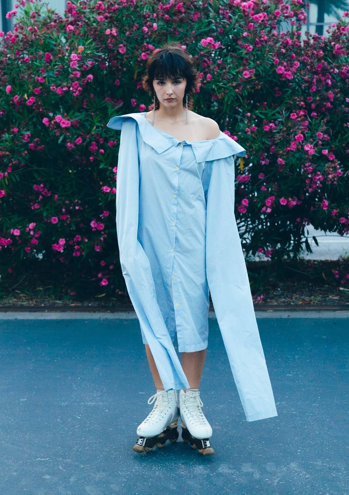 Una mujer con cabello rizado y corto, vestida con una camisa larga de color azul y patines de hielo, está de pie sobre una calle con un fondo de arbusto con flores rosadas.