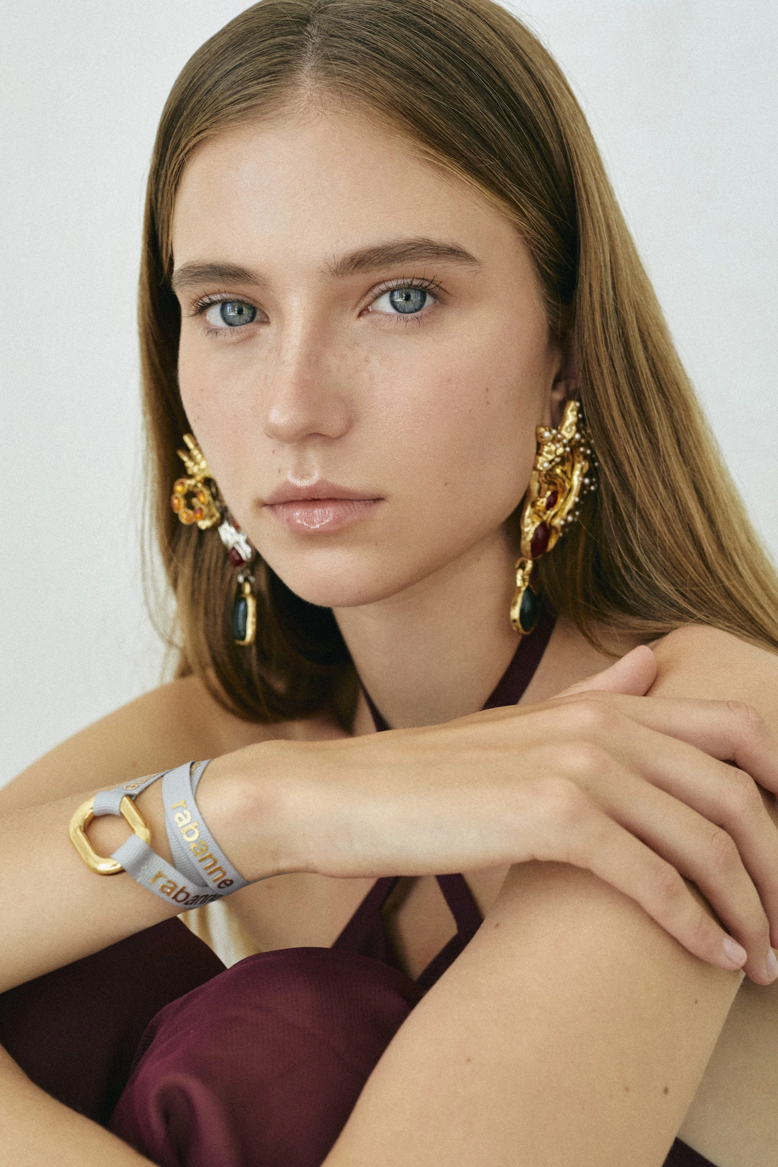 Retrato de una joven con cabello rubio, ojos azules, usando aretes grandes y un brazalete en la muñeca izquierda, en un fondo sencillo.