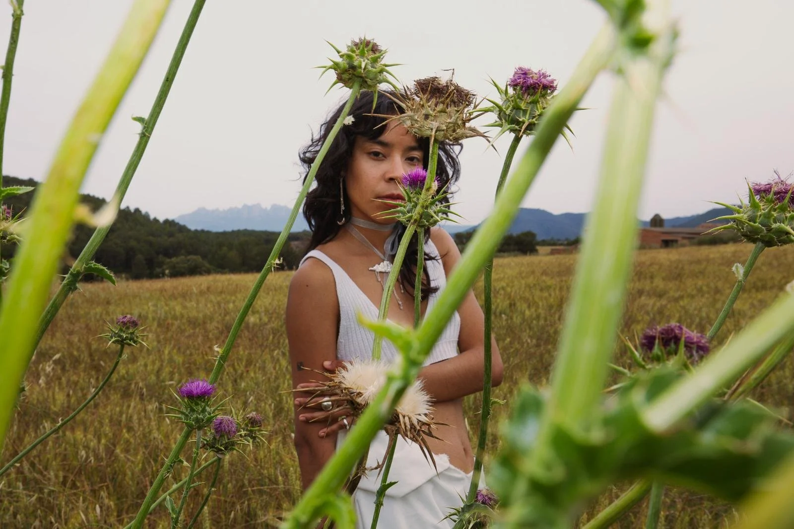 Mujer con cabello rizado oscuro y ropa blanca entre plantas de cardo con flores moradas en un campo con montañas al fondo.