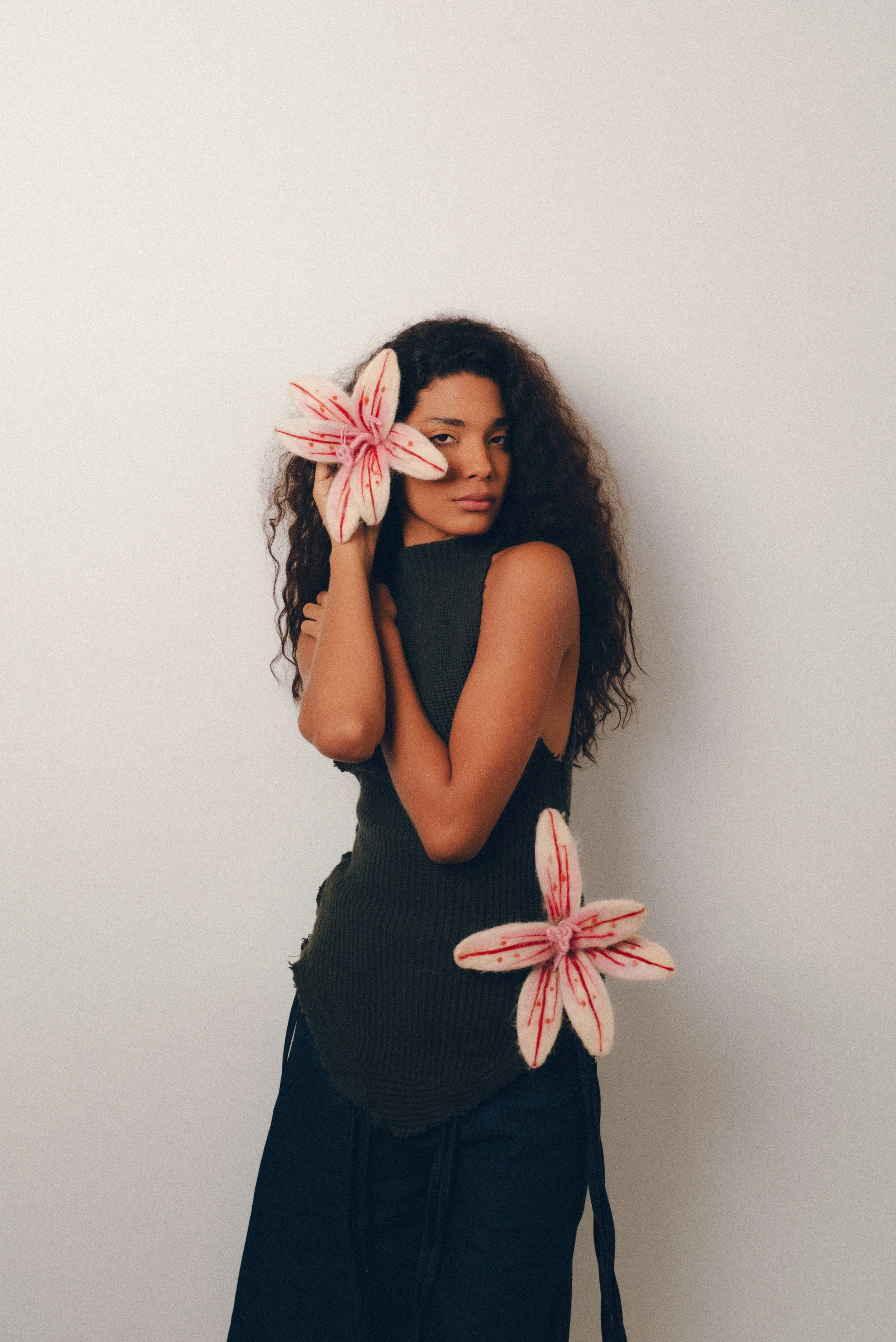 Una mujer con cabello rizado oscuro, vestida con un suéter negro y pantalones oscuros, sostiene dos flores de tela de color rosa con rojo y blanco, una cerca de su rostro y otra en su cintura, en un fondo blanco.