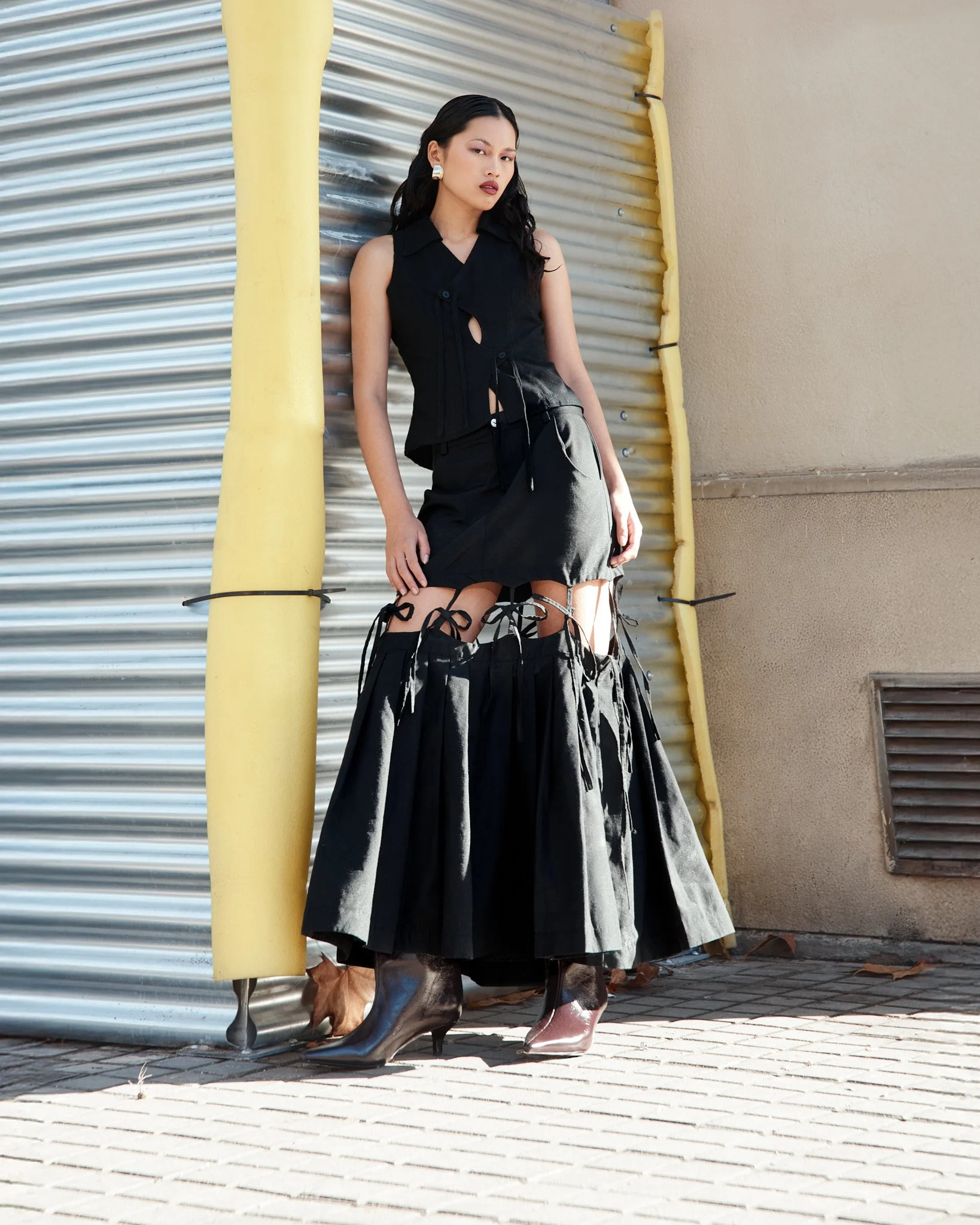 Mujer vestida con ropa negra y botas de tacón alto, posando al lado de una pared de metal y hormigón.