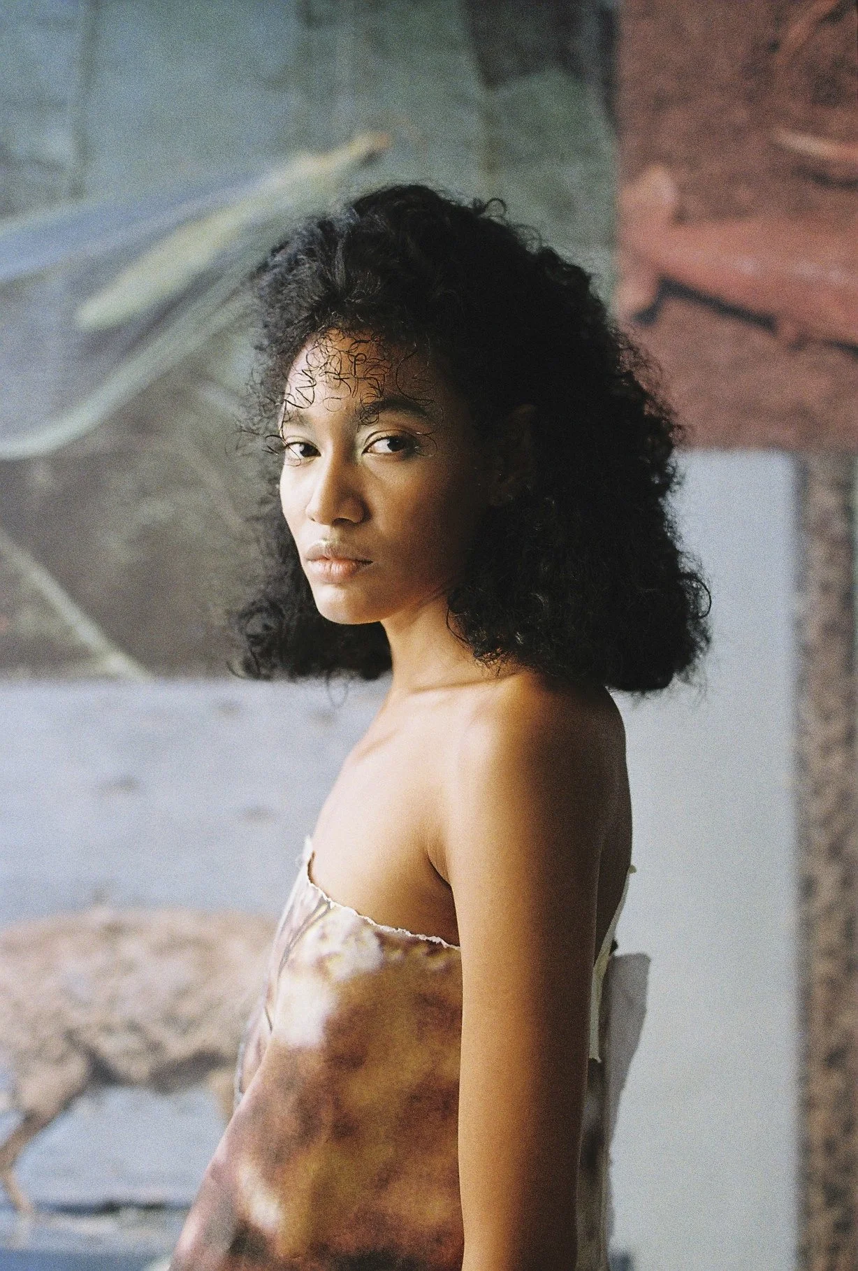 Retrato de una mujer joven con cabello rizado negro, piel morena, usando un vestido de seda en tonos tierras, en un ambiente con paredes de piedra y ladrillo.