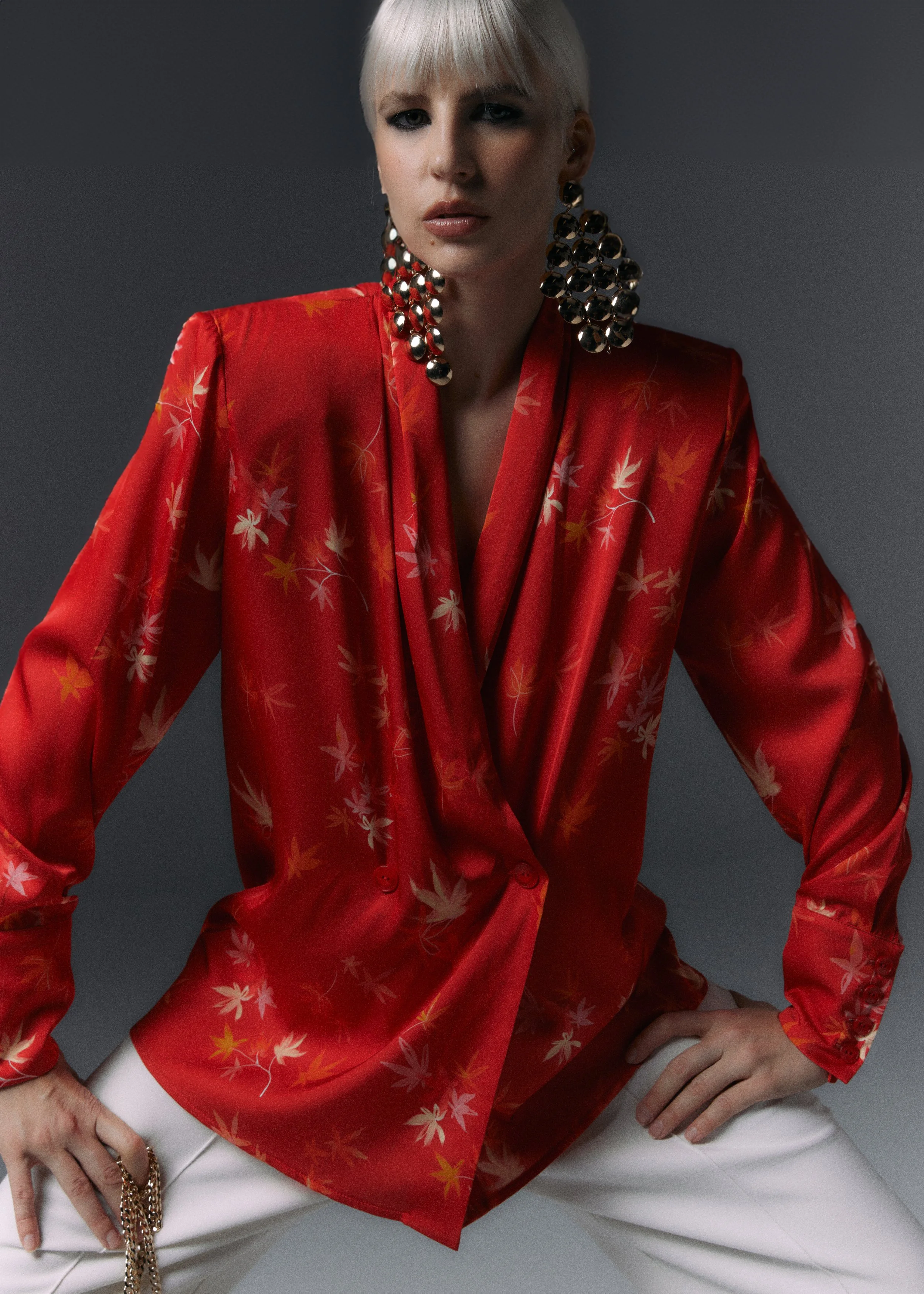 Mujer con cabello rubio y corto, vestida con un blazer rojo con estampado de hojas, pendientes grandes de bolas y pantalones blancos.