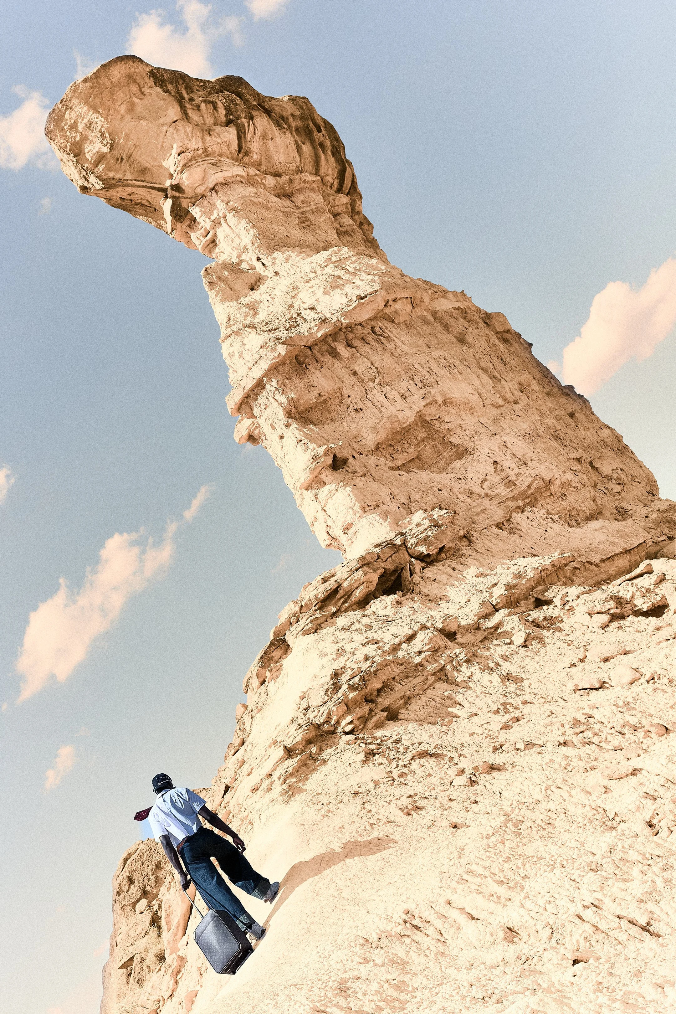 Persona con maleta caminando por un sendero en un paisaje árido y rocoso, con una formación de roca extensa en el fondo y cielo despejado con pocas nubes.