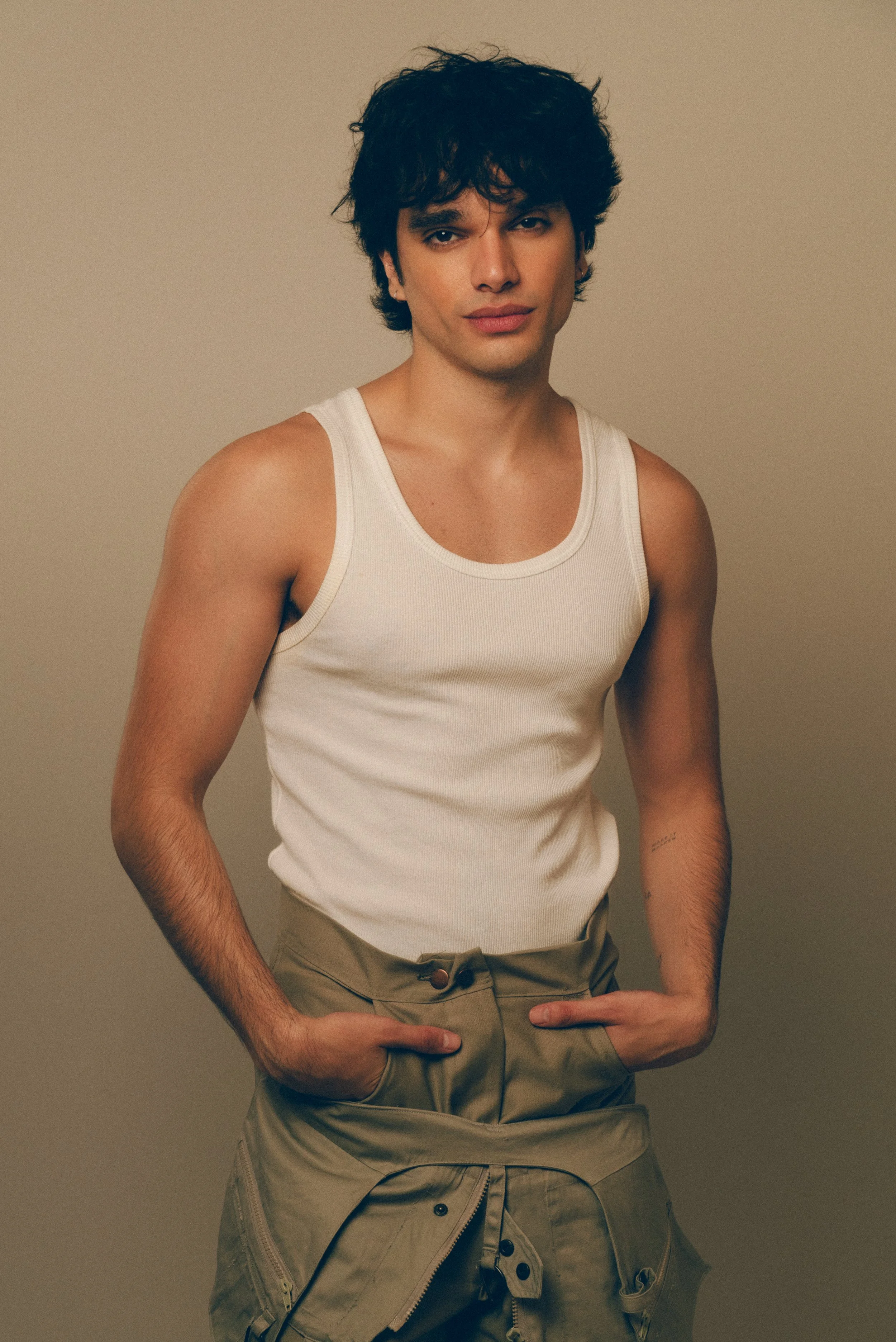 Joven con cabello oscuro, con camiseta sin mangas blanca y pantalones cortos beige, posando con las manos en los bolsillos y mirando hacia la cámara.