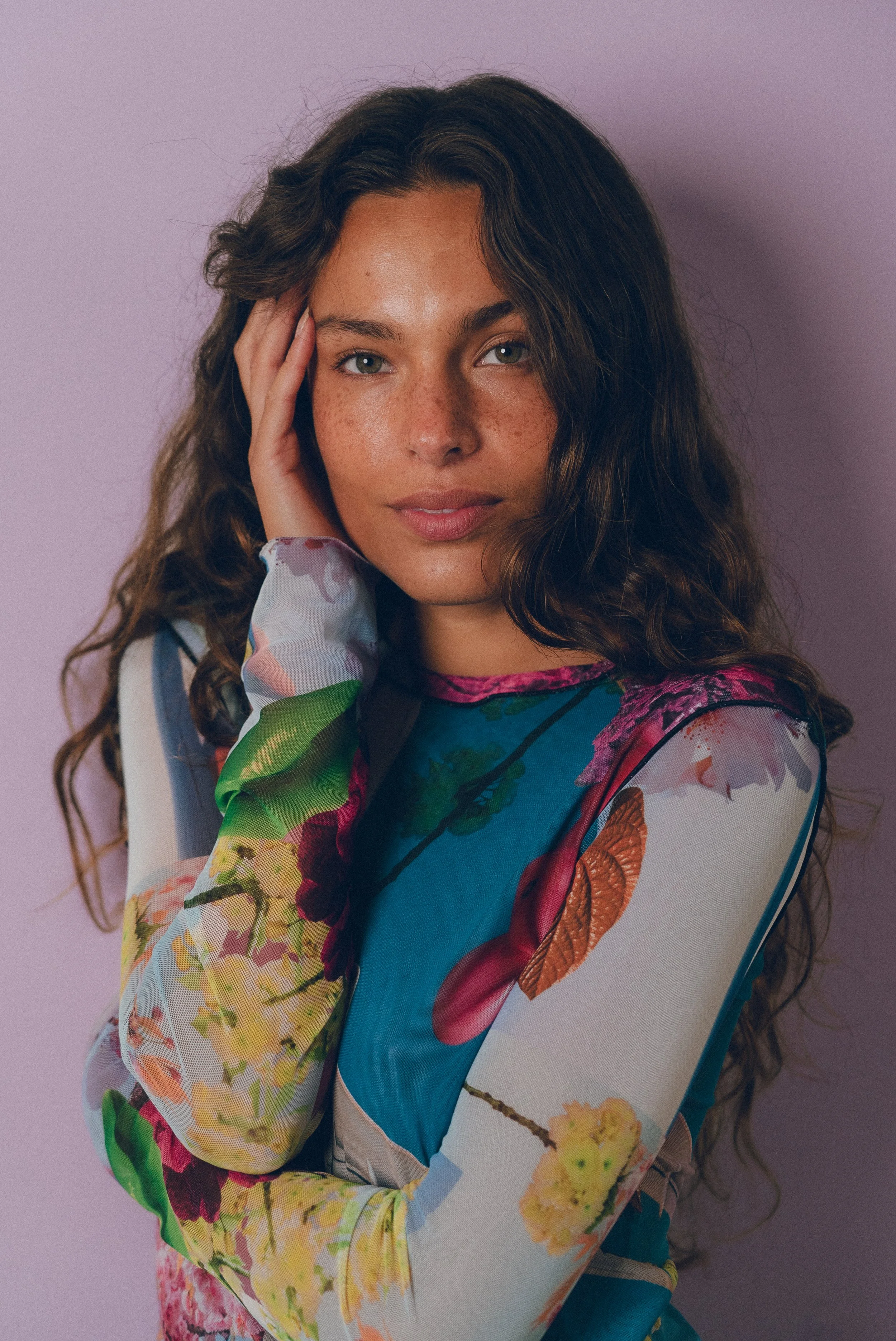 Retrato de una mujer con cabello rizado y piel bronceada, usando un vestido con estampado floral en tonos vibrantes, en un fondo lila.