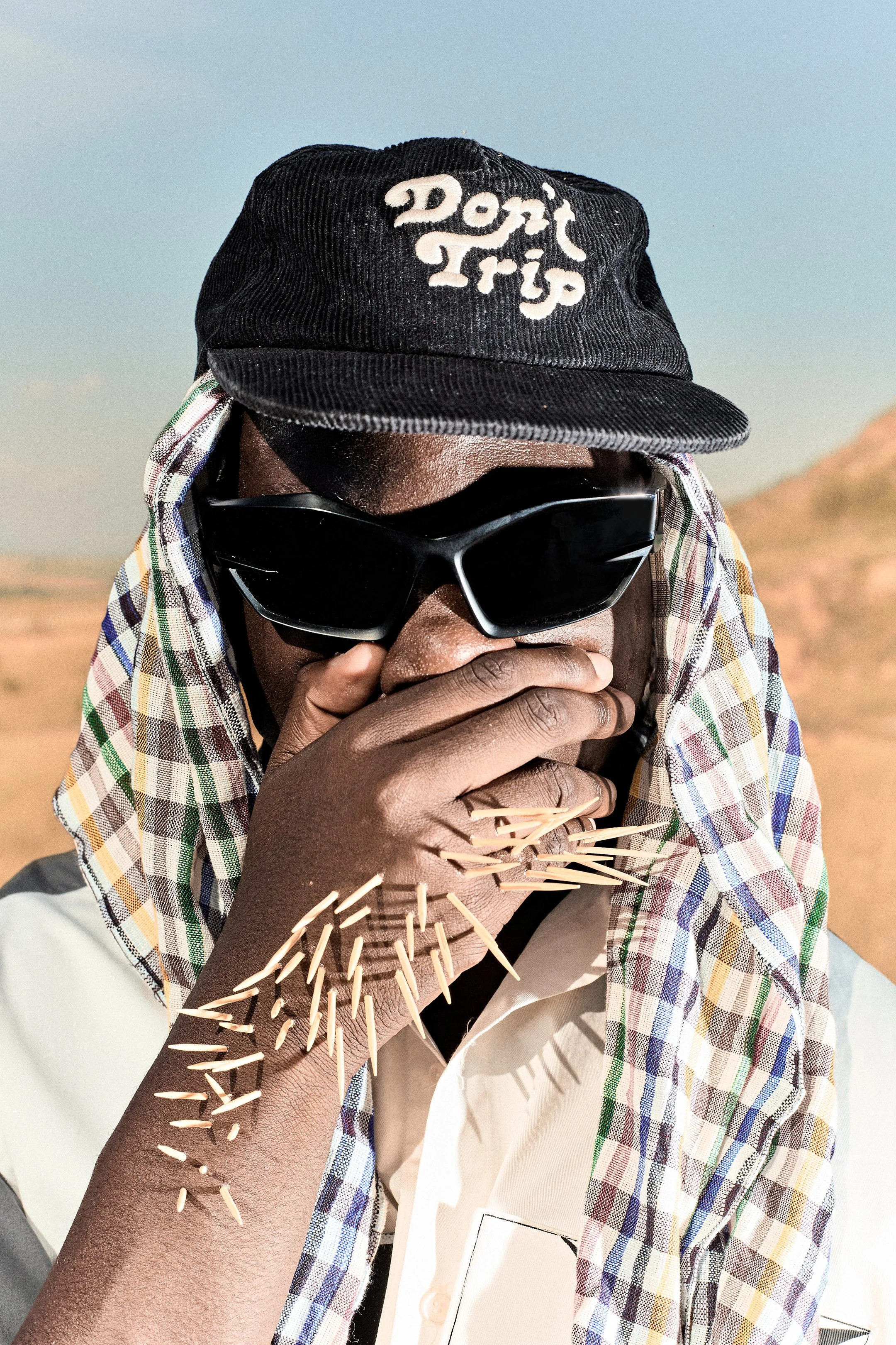 Persona con gafas de sol, sombrero y bufanda en un paisaje árido, sosteniendo una mano con horquillas bramante en la boca, con arena en la cara y brazo