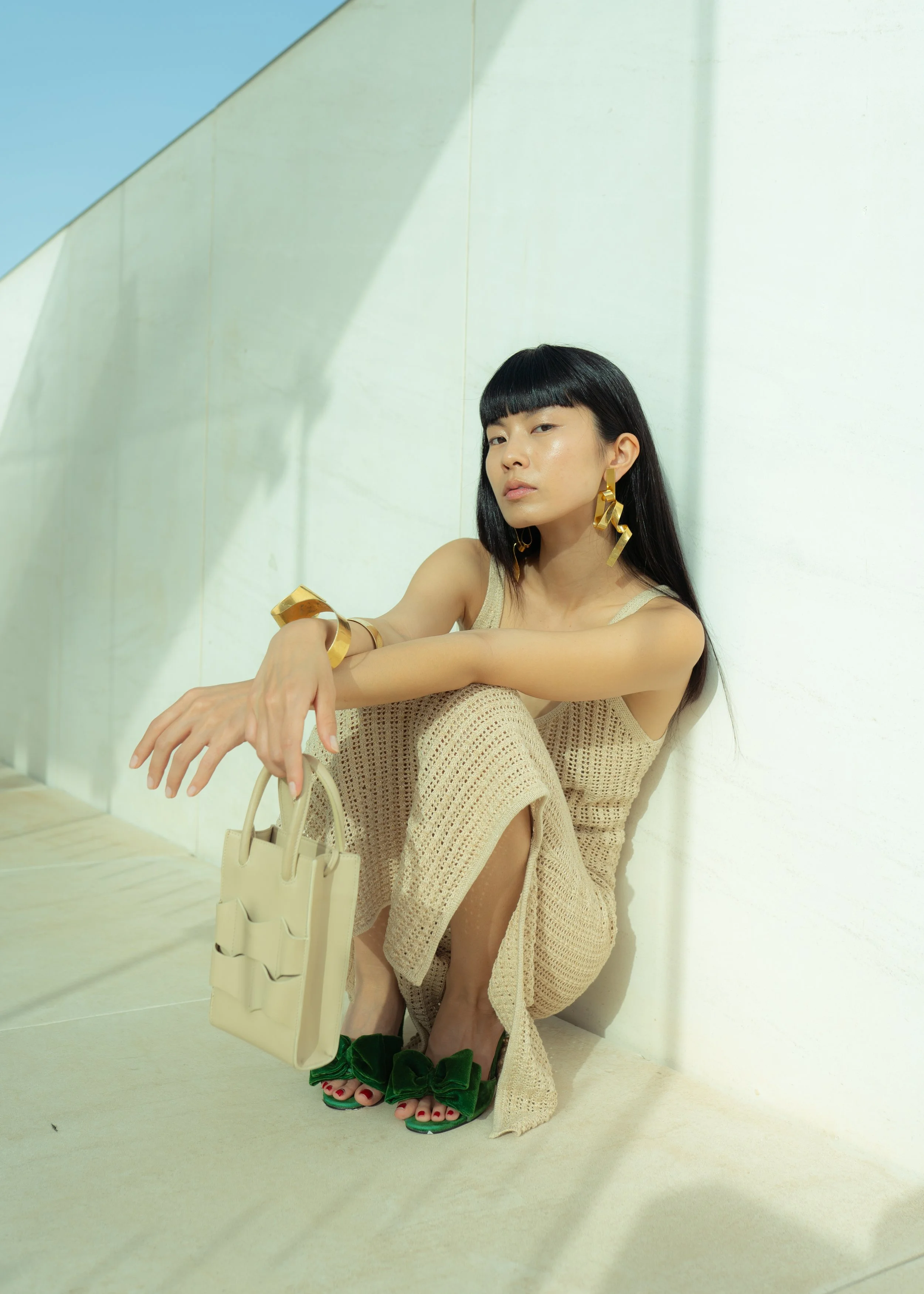Mujer sentada en el piso, vestida con ropa de crochet beige, con accesorios de oro, sosteniendo una bolsa beige, con sandalias verdes con lazo
