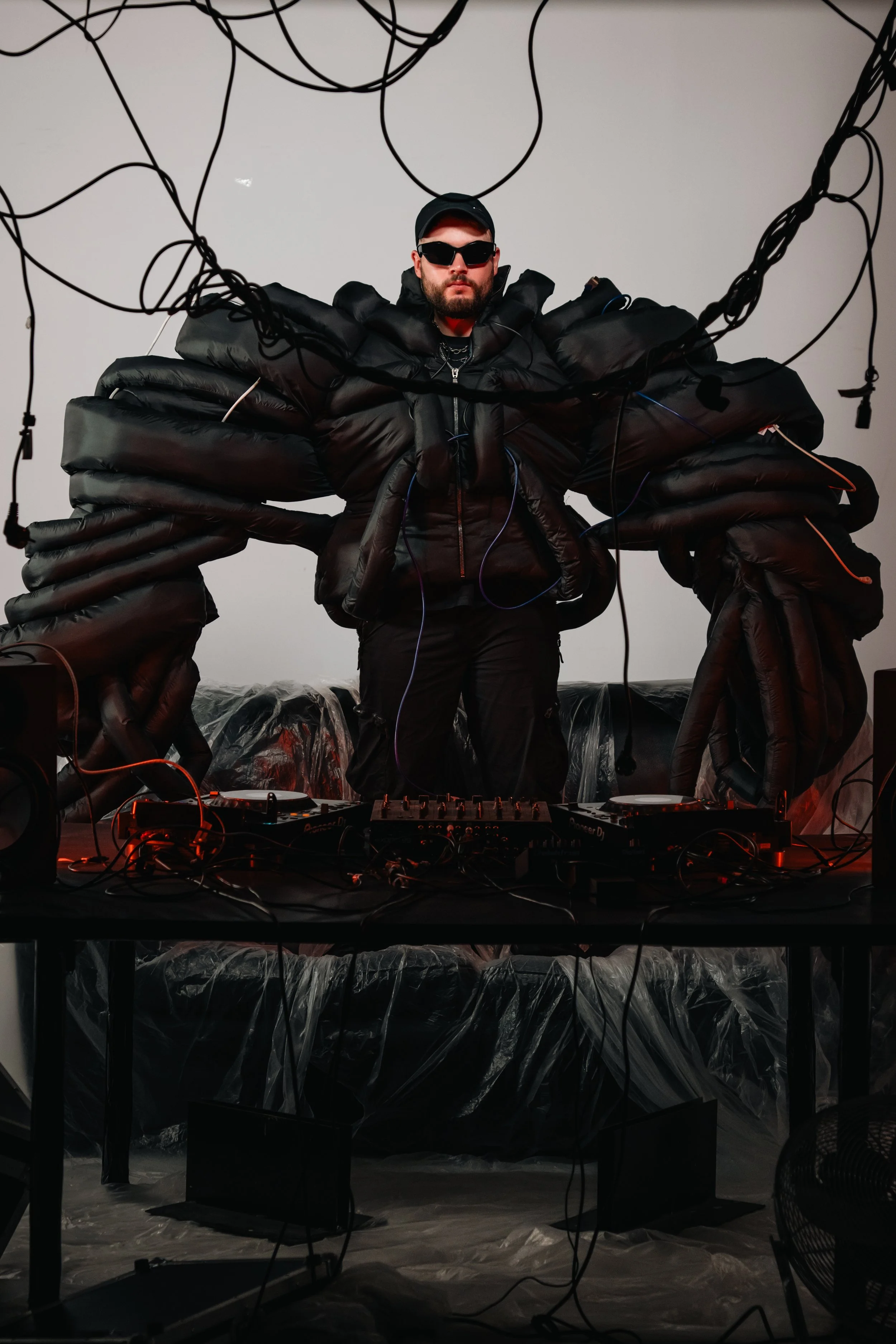 Una persona con gafas de sol y ropa negra en medio de una instalación artística de objetos inflados negros y cables, con equipo de música en una mesa al frente.