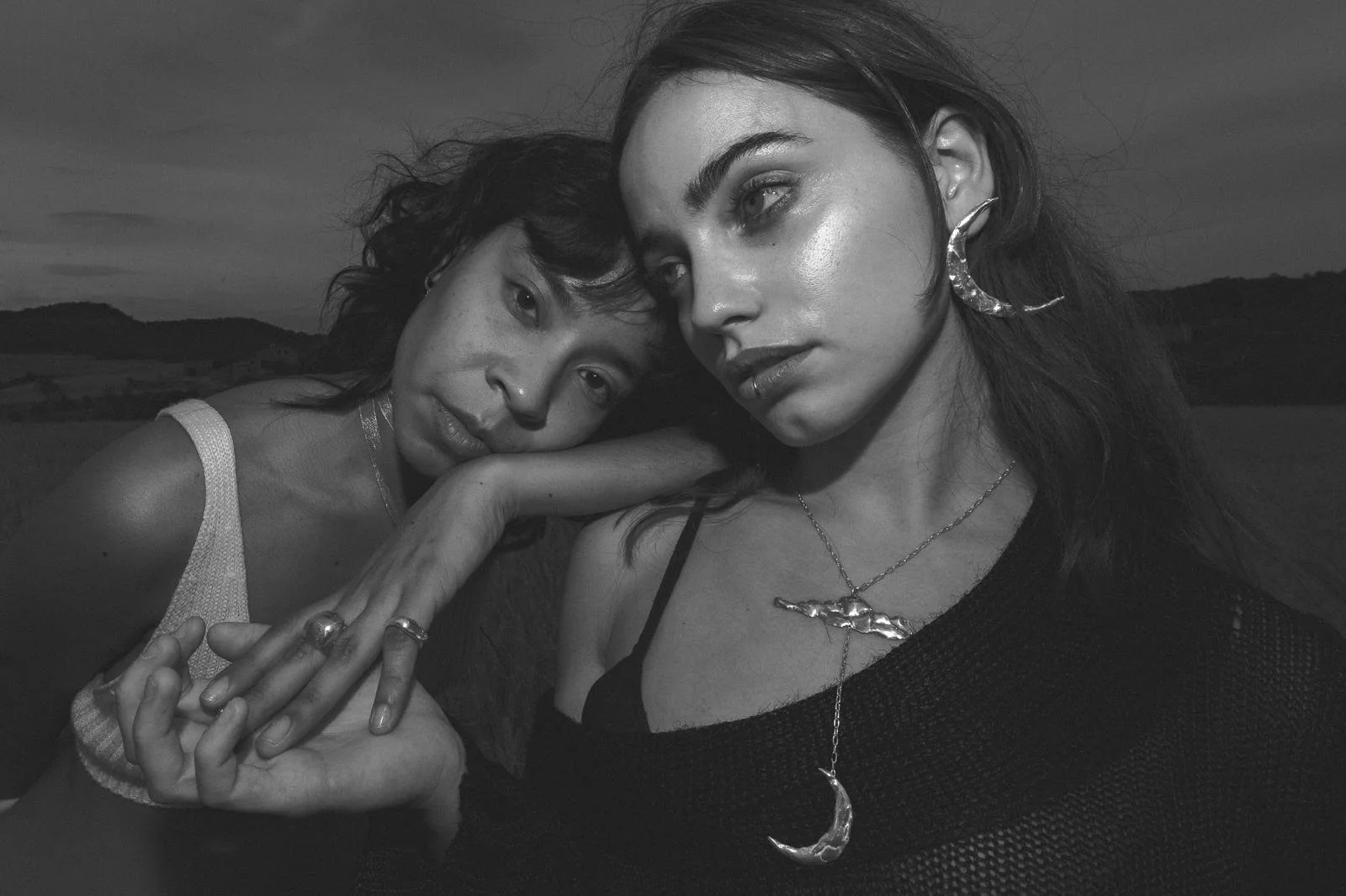 Dos mujeres con joyería en un paisaje al atardecer, una con cabello rizado y la otra con cabello liso, posando juntas en un retrato en blanco y negro.