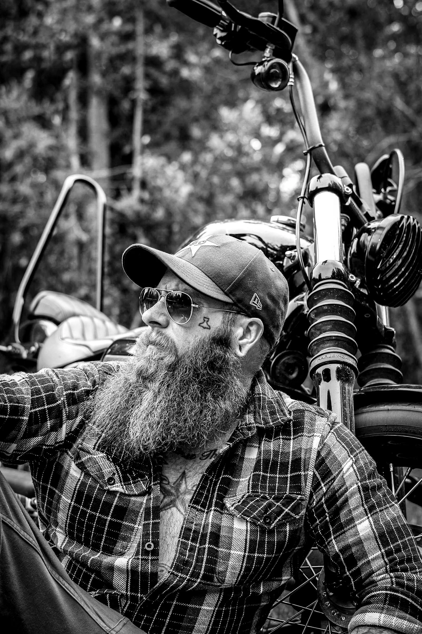 A bearded man wearing sunglasses, a cap, and a plaid shirt, leaning against a motorcycle in a forested area.