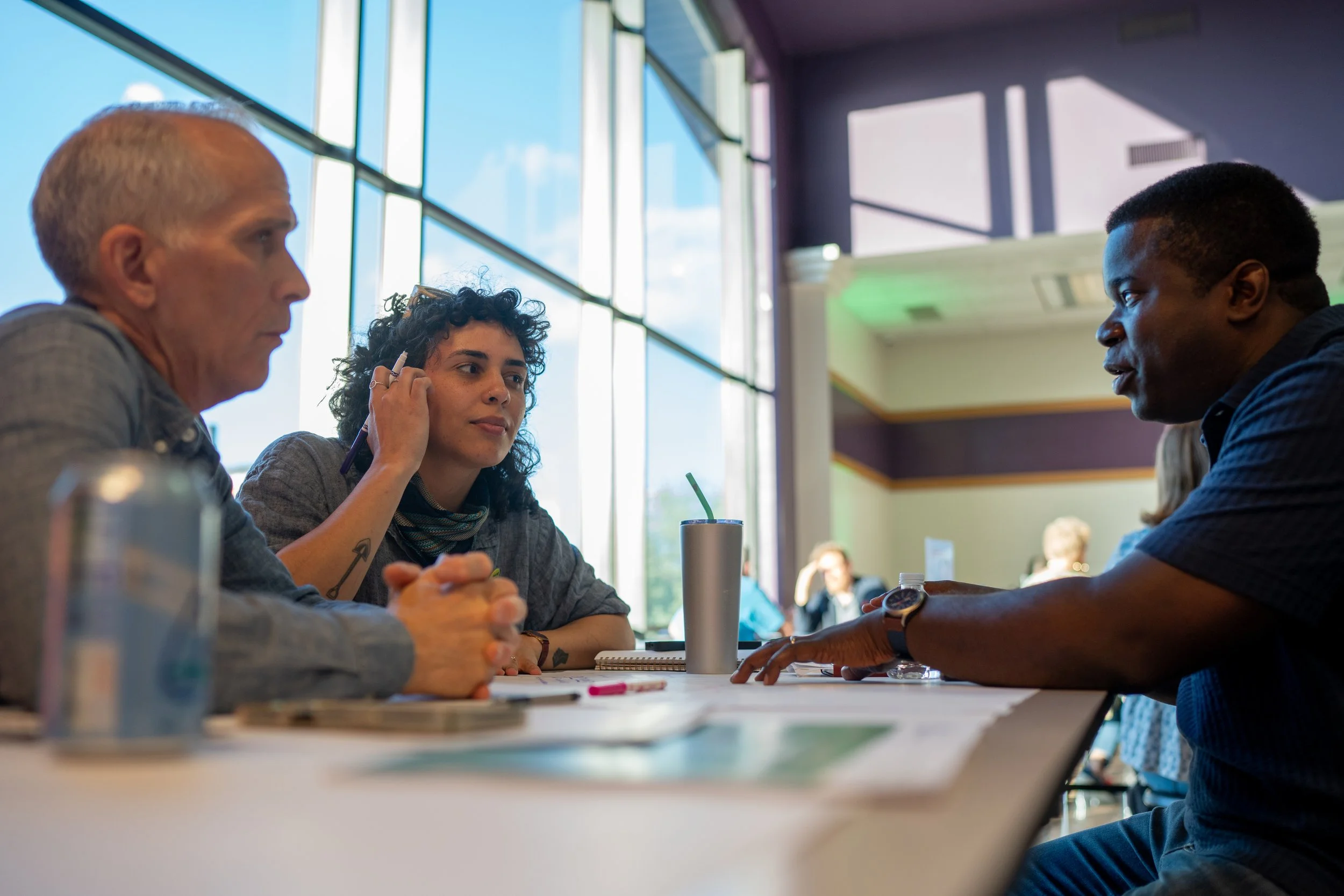 Three people sitting at a table engaged in discussion inside a well-lit room with large windows.