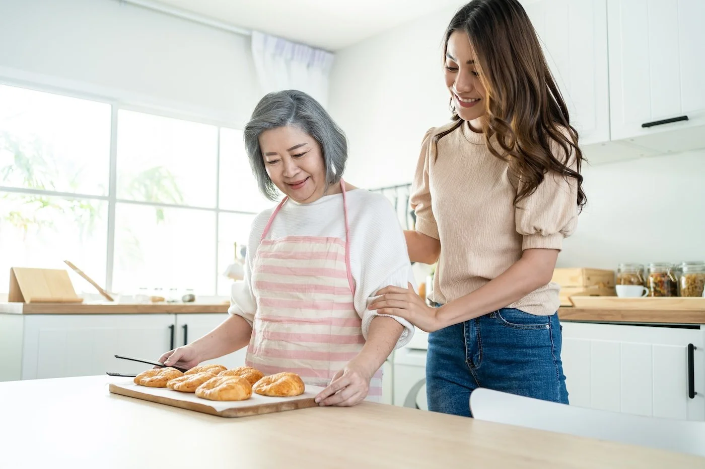 A young woman helps an elderly woman with baked pastries in a bright kitchen.