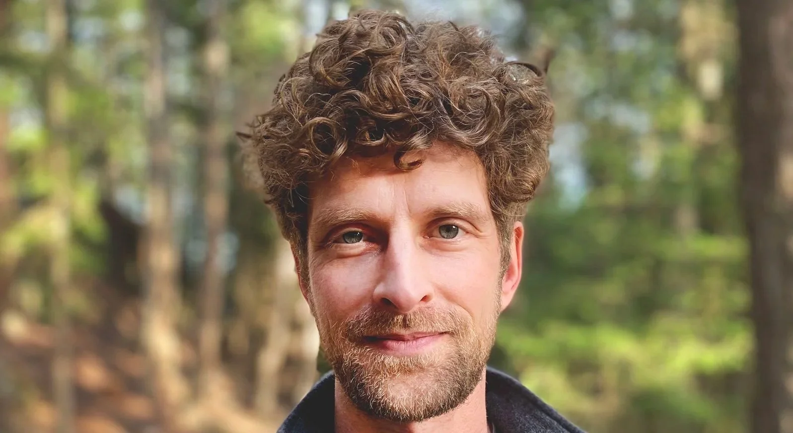 Close-up of a man with curly brown hair and a beard, smiling outdoors in a forest with trees and greenery in the background.