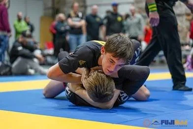 Two young boys are practicing Brazilian Jiu-Jitsu on a blue mat, with one boy on top in a dominant position, pinning the other.